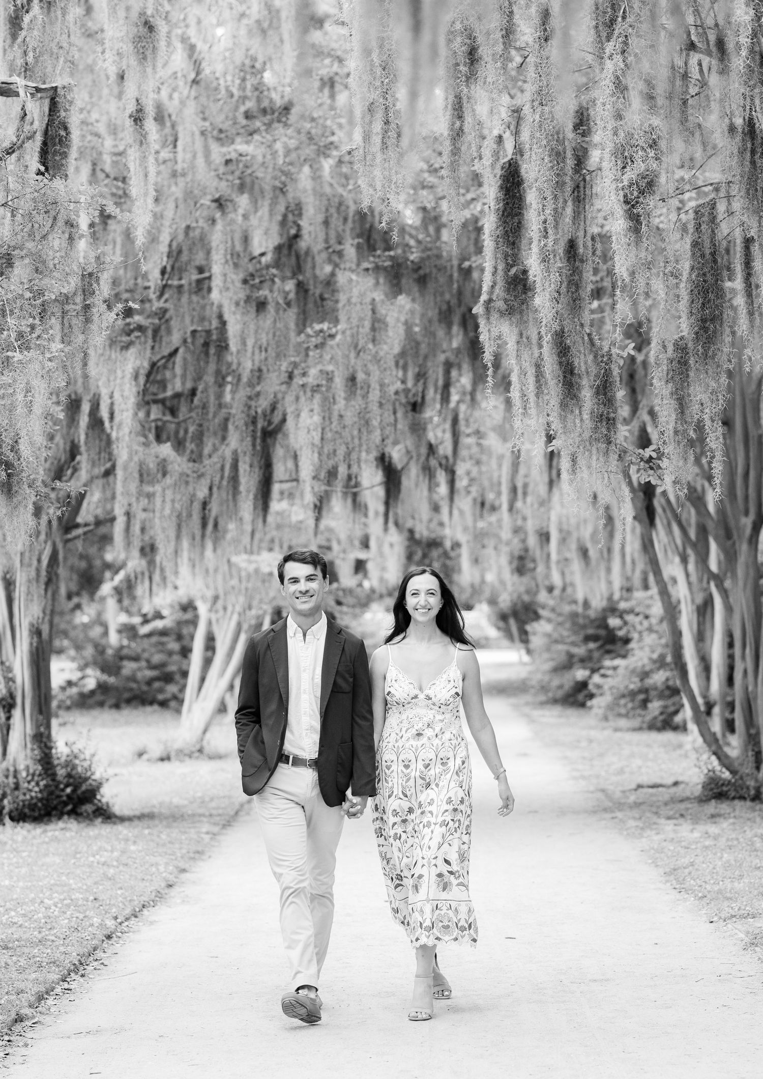 Black and white photo of a couple walking hand in hand down a tree-lined path draped with Spanish moss.