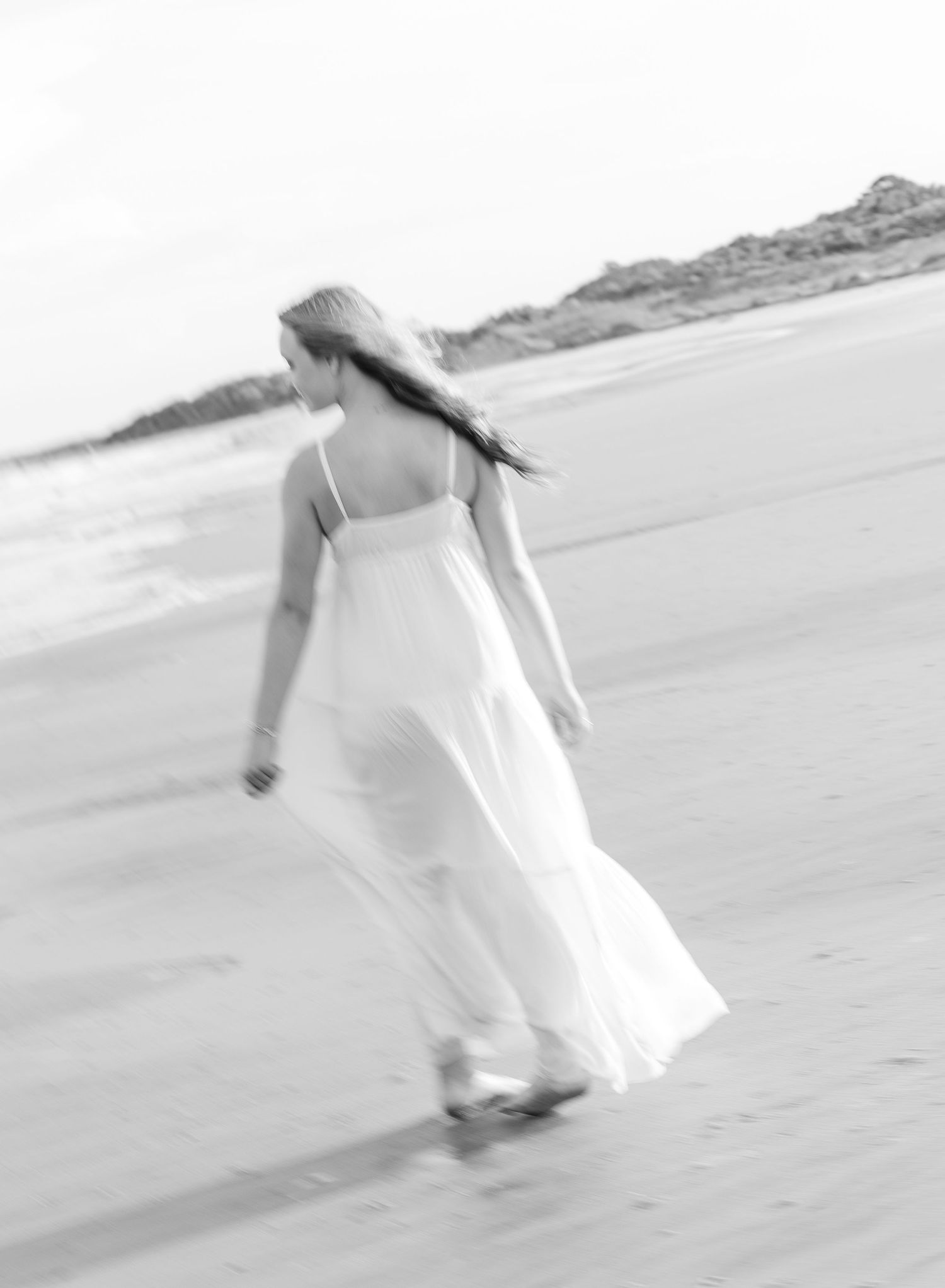 A black and white photo captures a flowing white dress moving in the ocean breeze on a sandy beach.