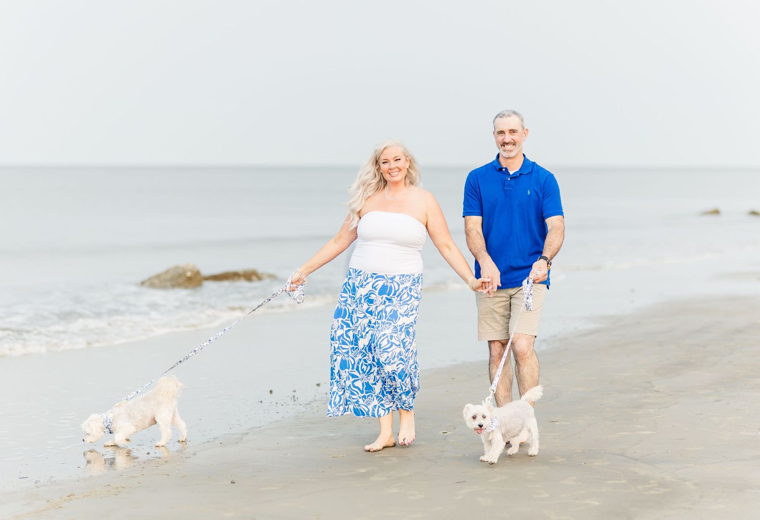 Two people walk their small white dogs along the beach shore wearing blue and white summer outfits.