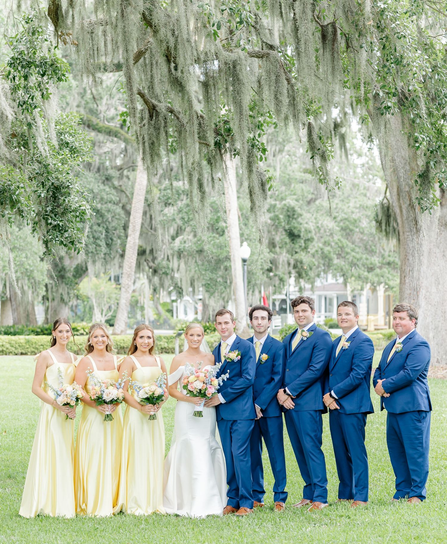 Wedding party in navy blue suits and pastel yellow bridesmaid dresses pose outdoors under Spanish moss draped trees.