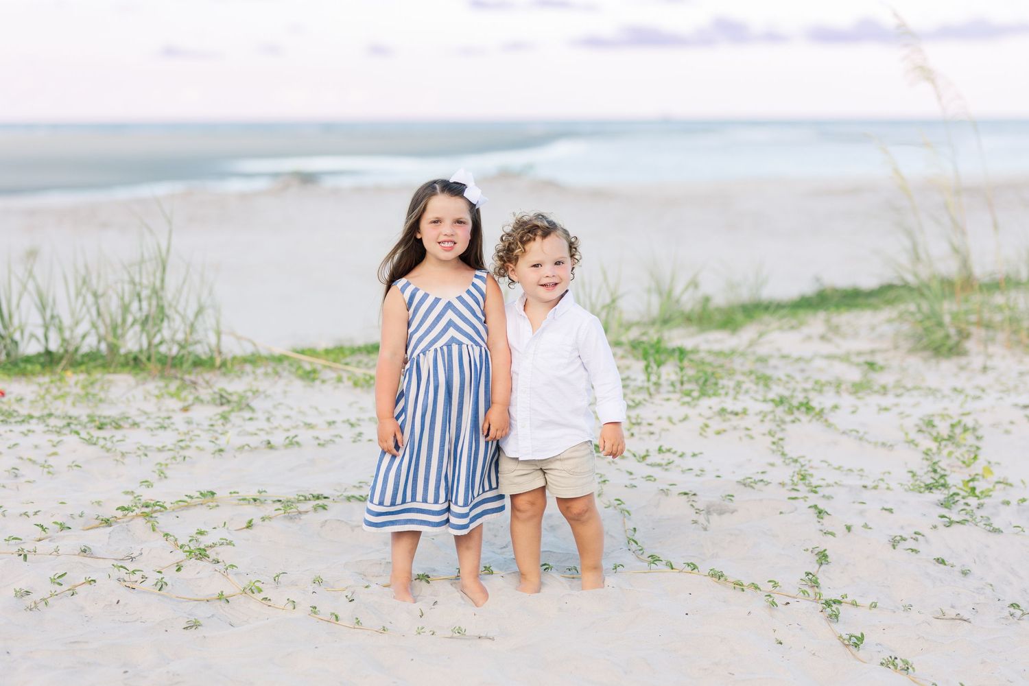 Two young children stand together smiling on a sandy beach with ocean waves in the background.