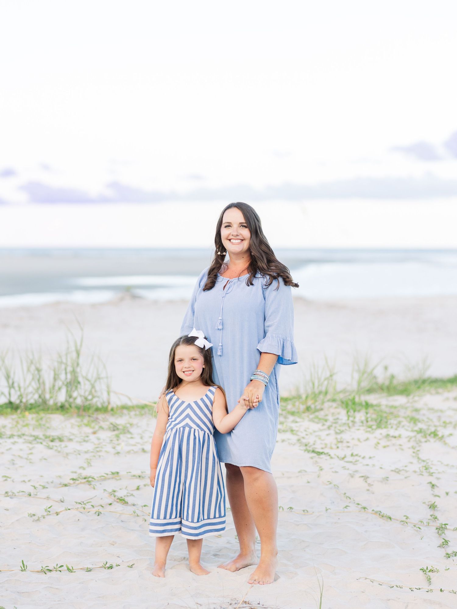 A parent and child wearing coordinating blue and white outfits stand together barefoot on a sandy beach.