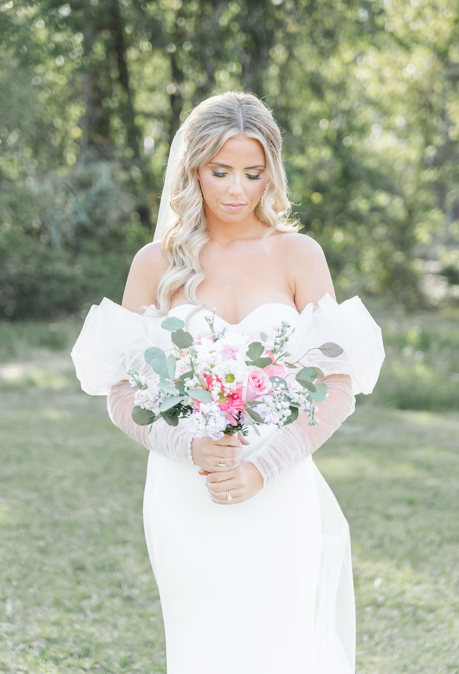 Bride in off-shoulder white gown holds romantic pink and eucalyptus bouquet in outdoor garden setting.