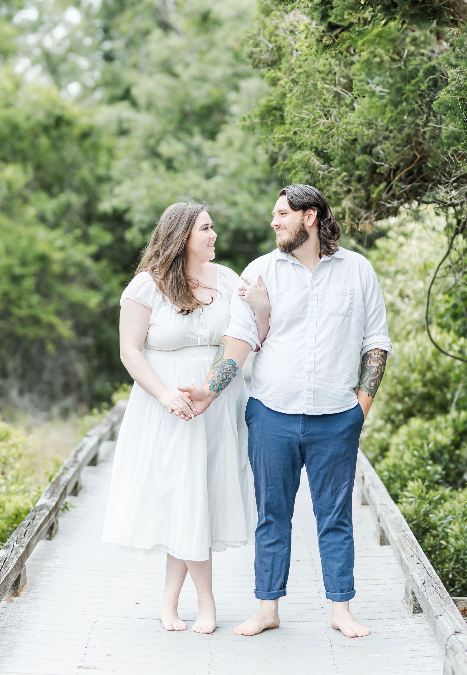 Couple shares loving glance while standing barefoot on wooden walkway.