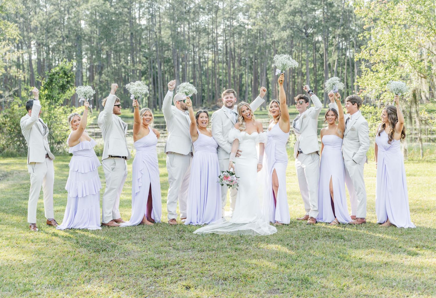 Wedding party in white and lilac dresses poses joyfully on a sunny lawn with forest backdrop.
