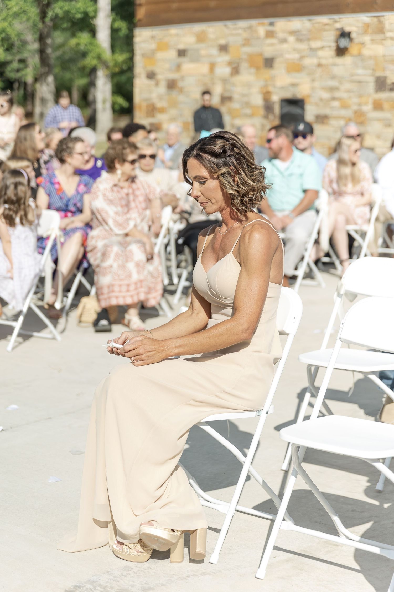 Wedding guest in neutral dress seated on white folding chair during outdoor ceremony.