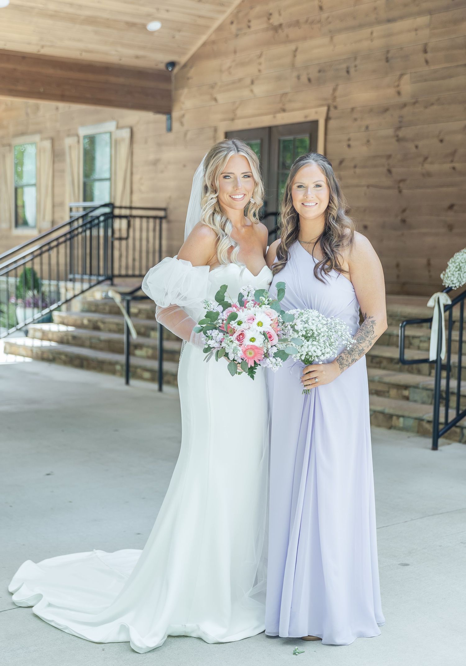 A bride in a white gown and bridesmaid in lavender dress pose with floral bouquets on a rustic wooden porch.