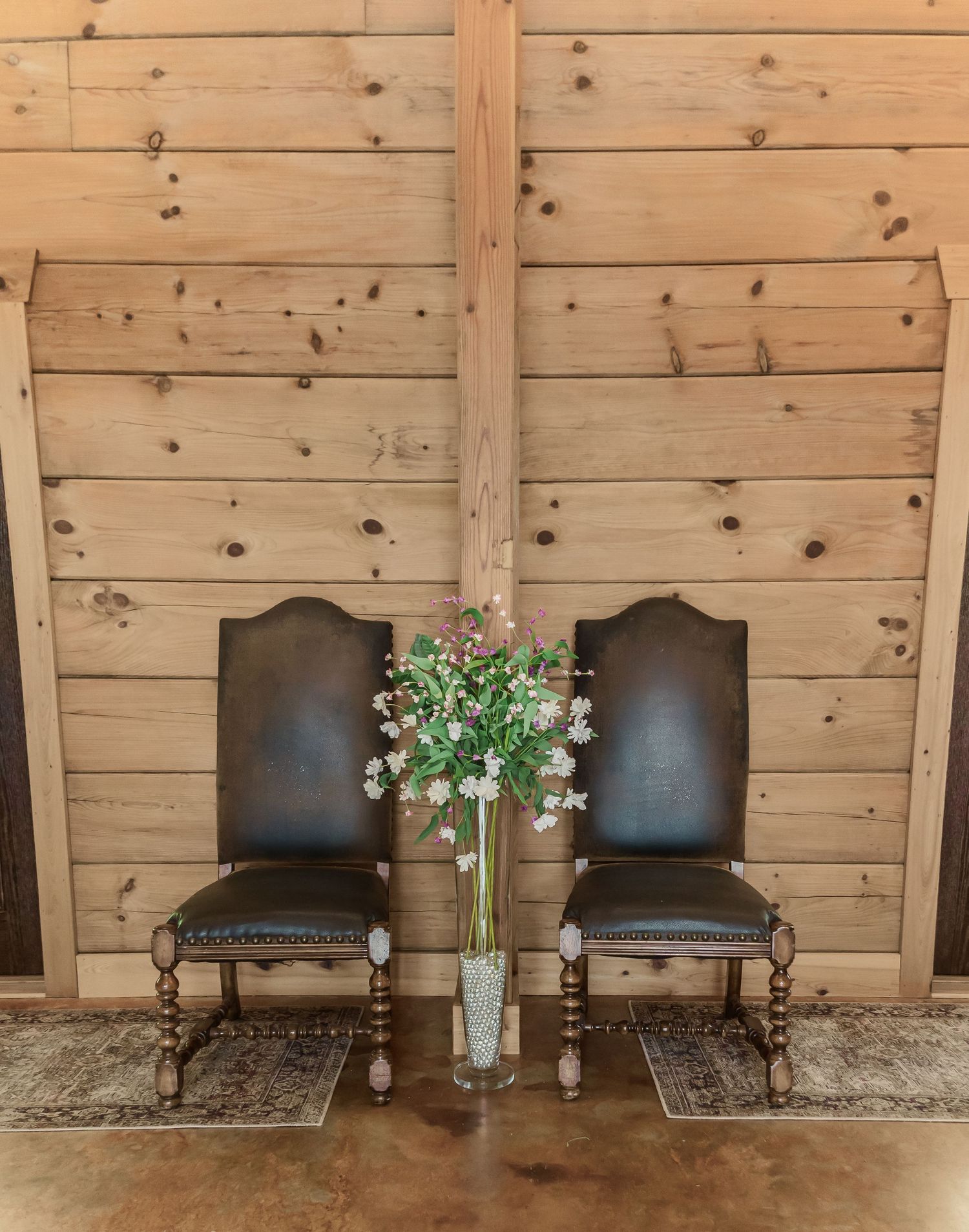 Two vintage leather chairs against wooden wall with tall floral arrangement between them.