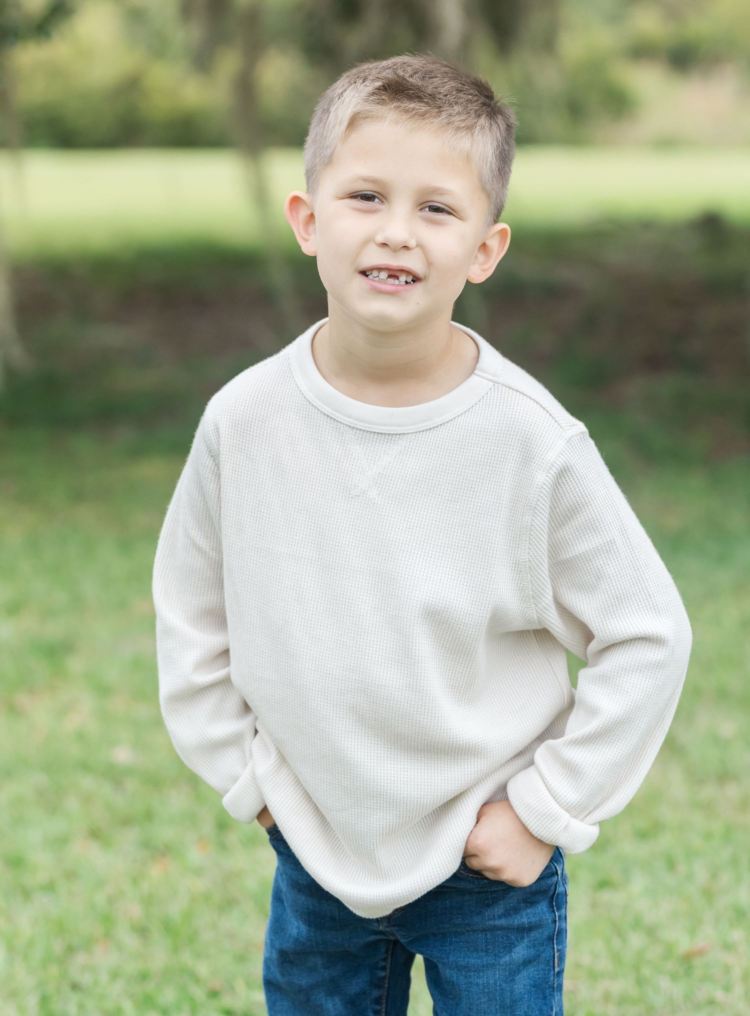 Young child in white sweater and jeans poses playfully for the camera outdoors.