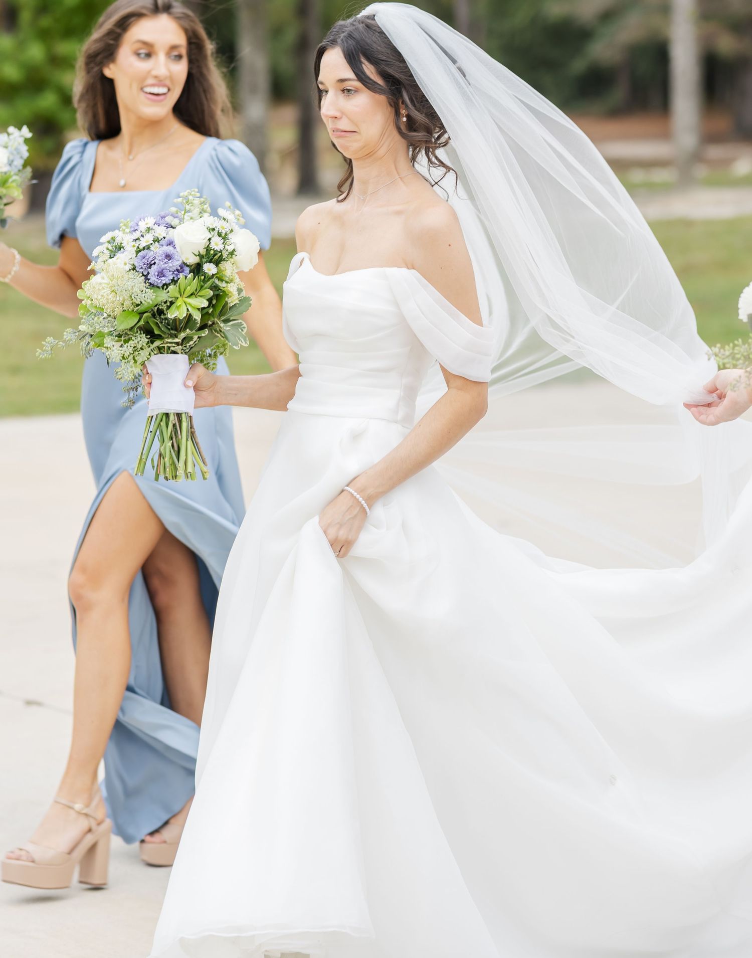 Bride in white gown with flowing veil stands beside bridesmaid in light blue dress.