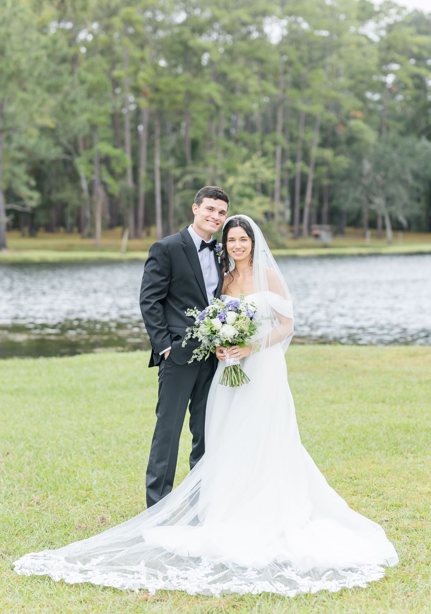A romantic outdoor wedding scene shows a couple in formal attire by a lake surrounded by tall pine trees.