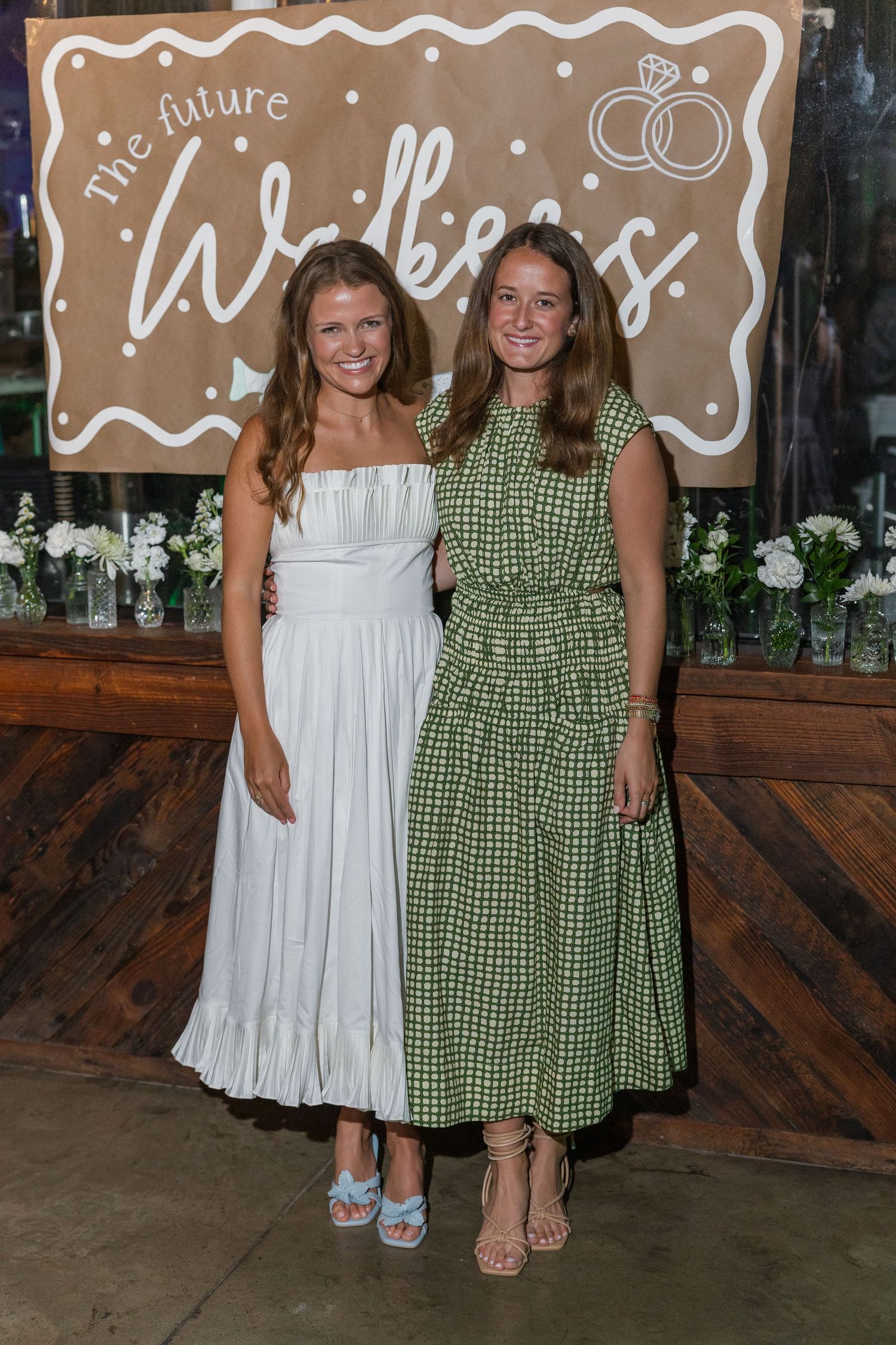 Two women in white and green dresses pose before The Future Walkers engagement party sign.
