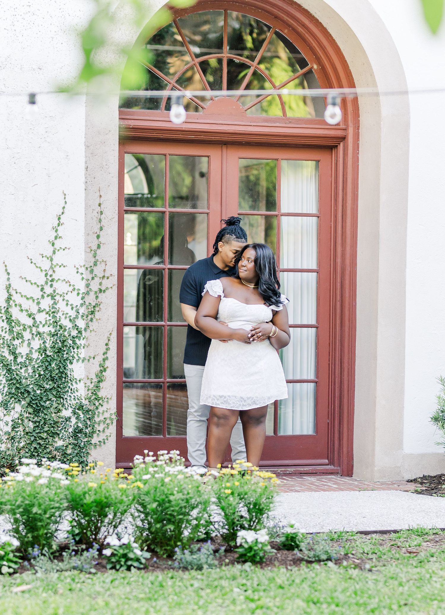 Couple embracing in front of elegant red French doors surrounded by lush garden flowers during an outdoor engagement session.