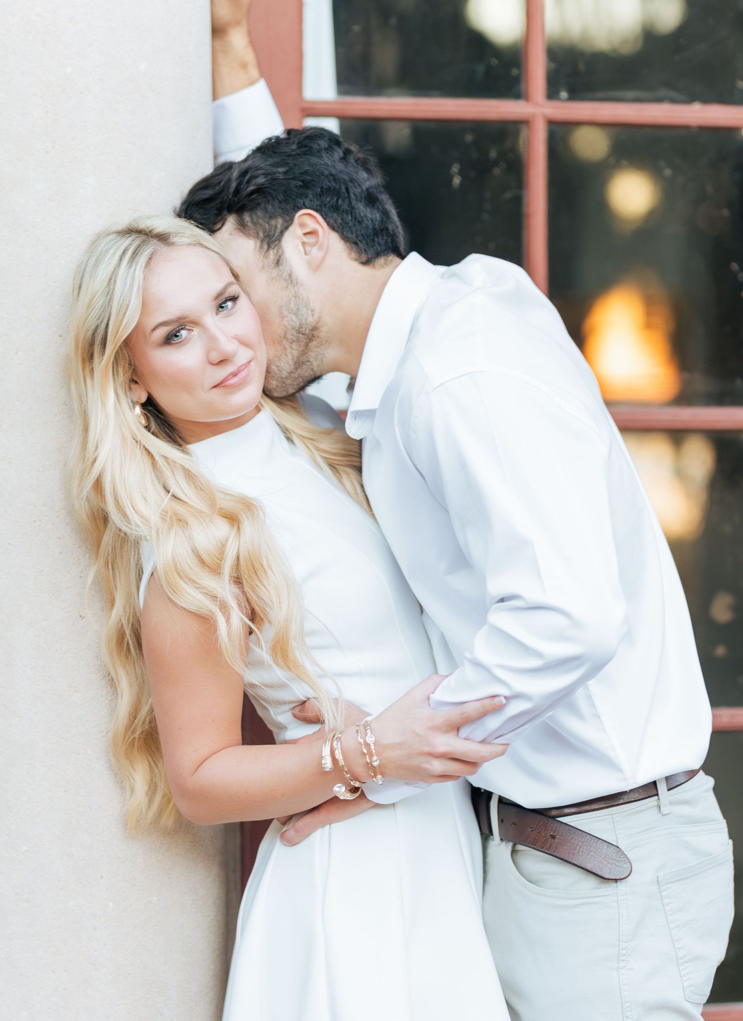 A couple in white outfits share a romantic moment, man kissing woman's temple near a rustic window.