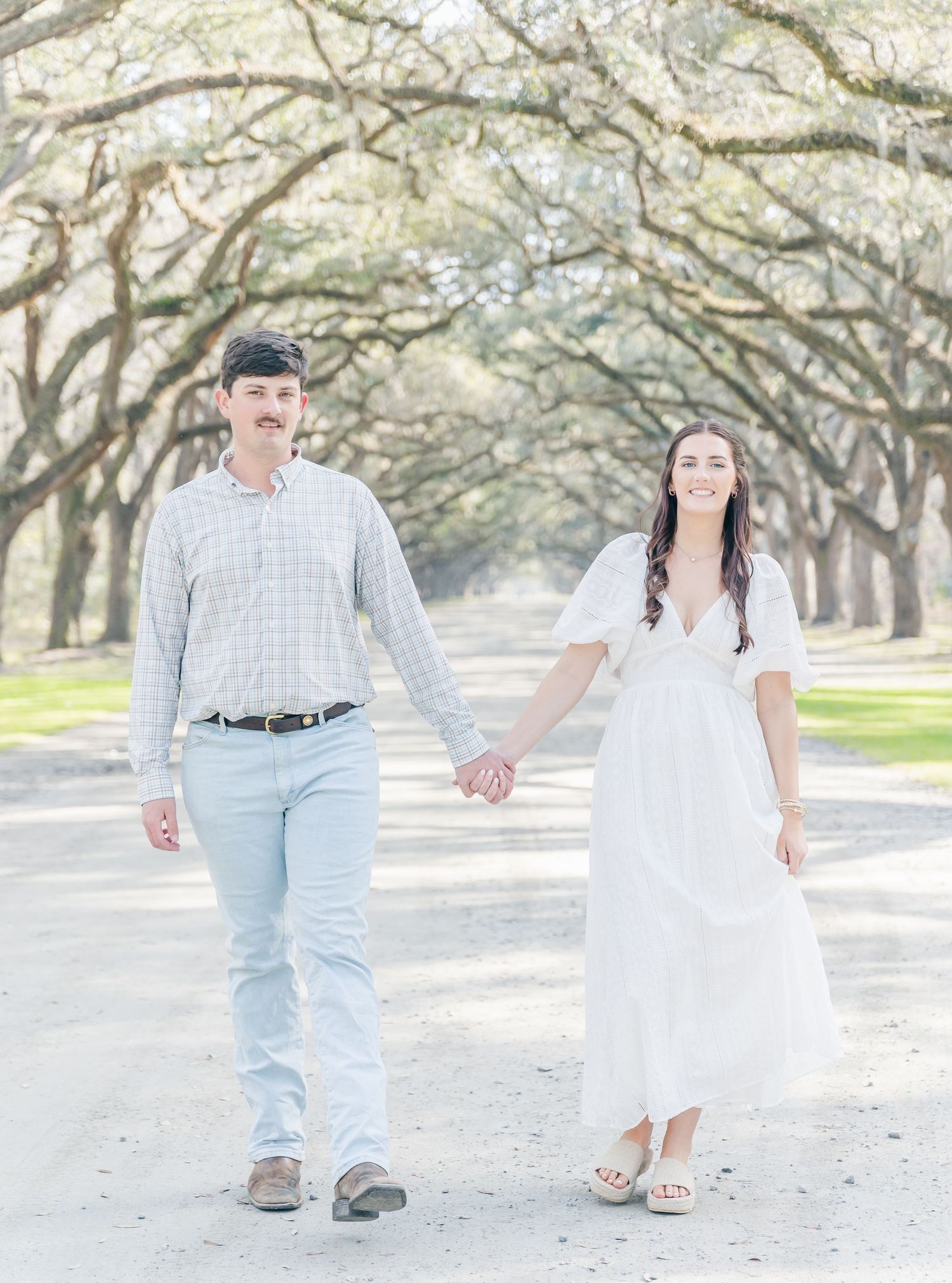A couple walks hand in hand down a tree-lined path, with the woman in a white dress and man in light casual attire.