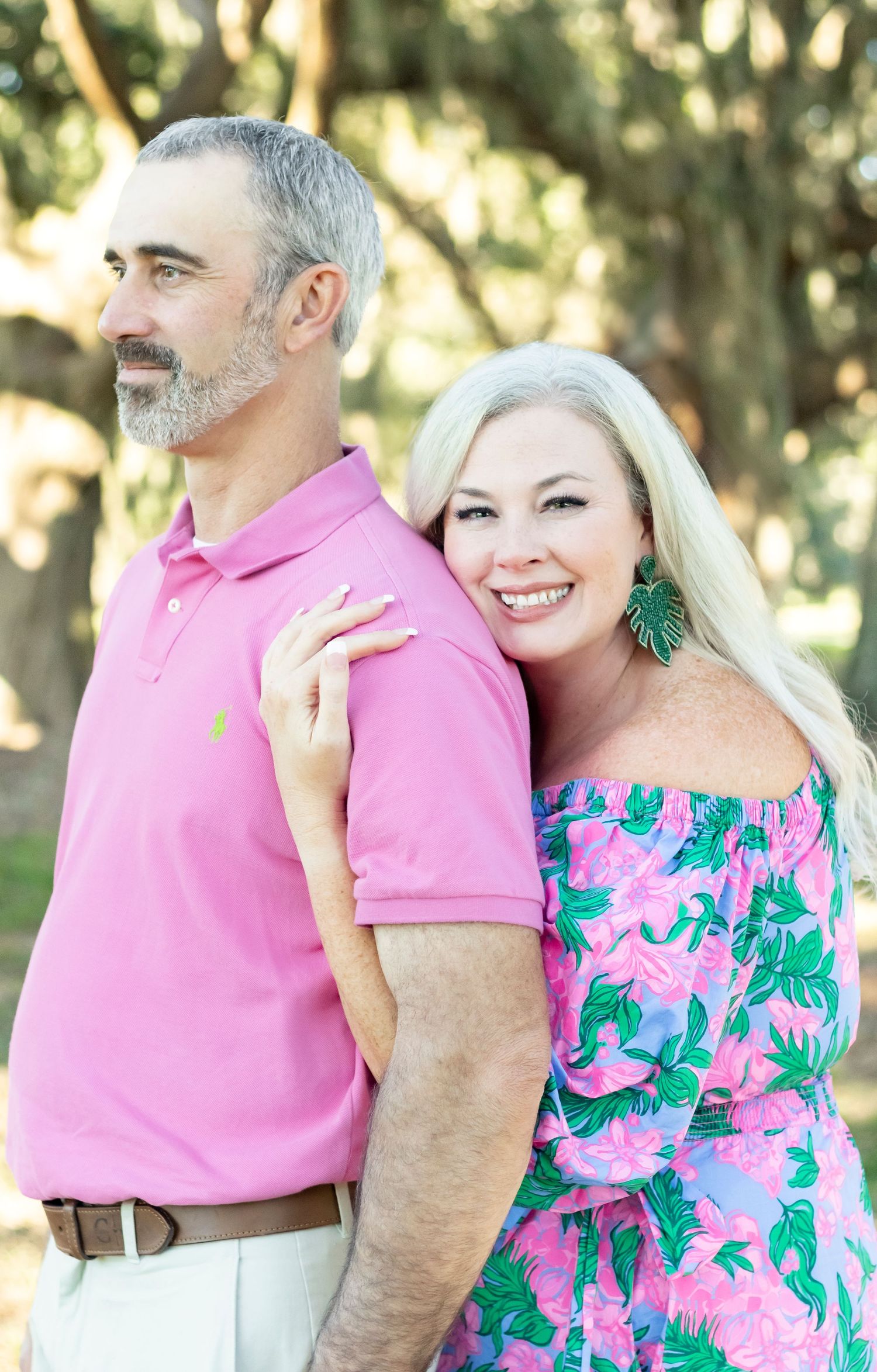 Couple poses outdoors in coordinated pink and floral outfits against a sunlit garden backdrop.