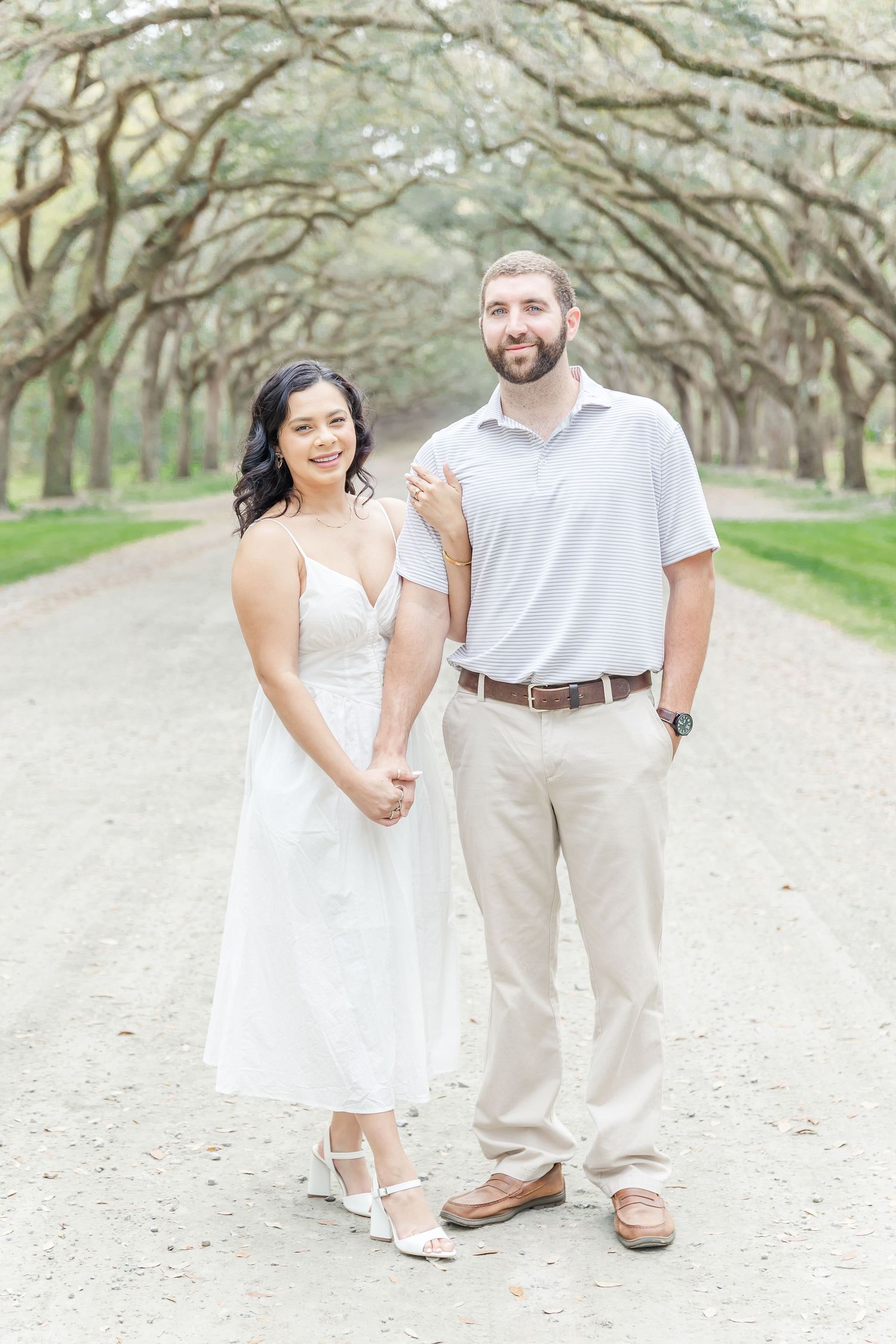 Elegant couple in white and khaki stand together on oak-lined dirt road.