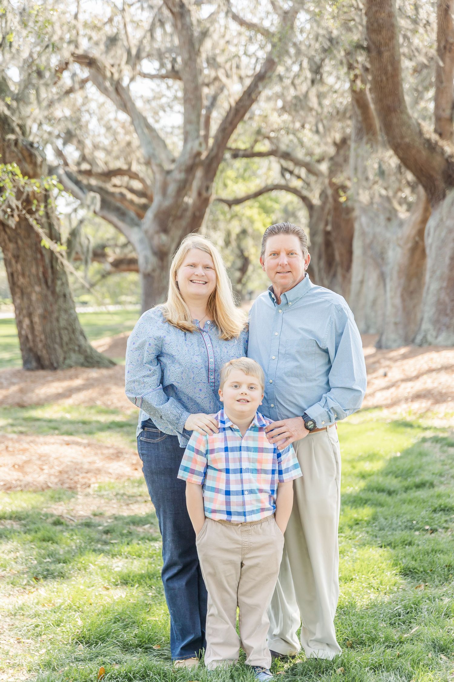 Family of three wearing coordinated blue outfits poses together under large oak trees in a park setting.