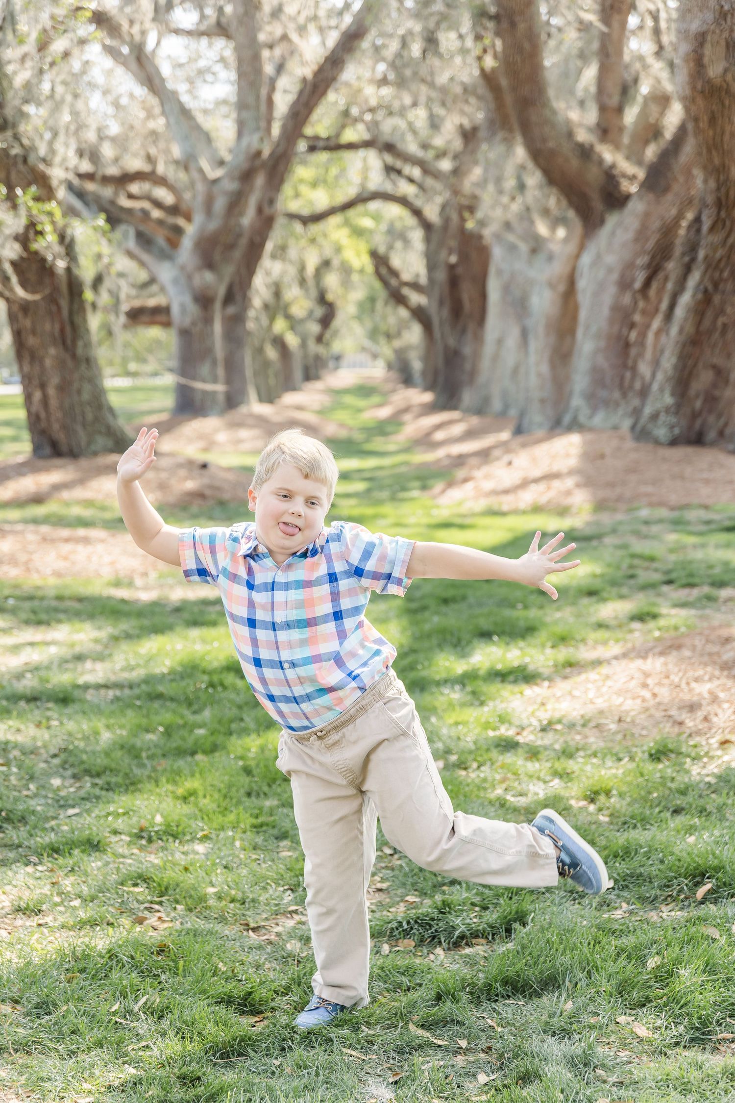 Energetic pose with arms outstretched on grass under tree-lined path.