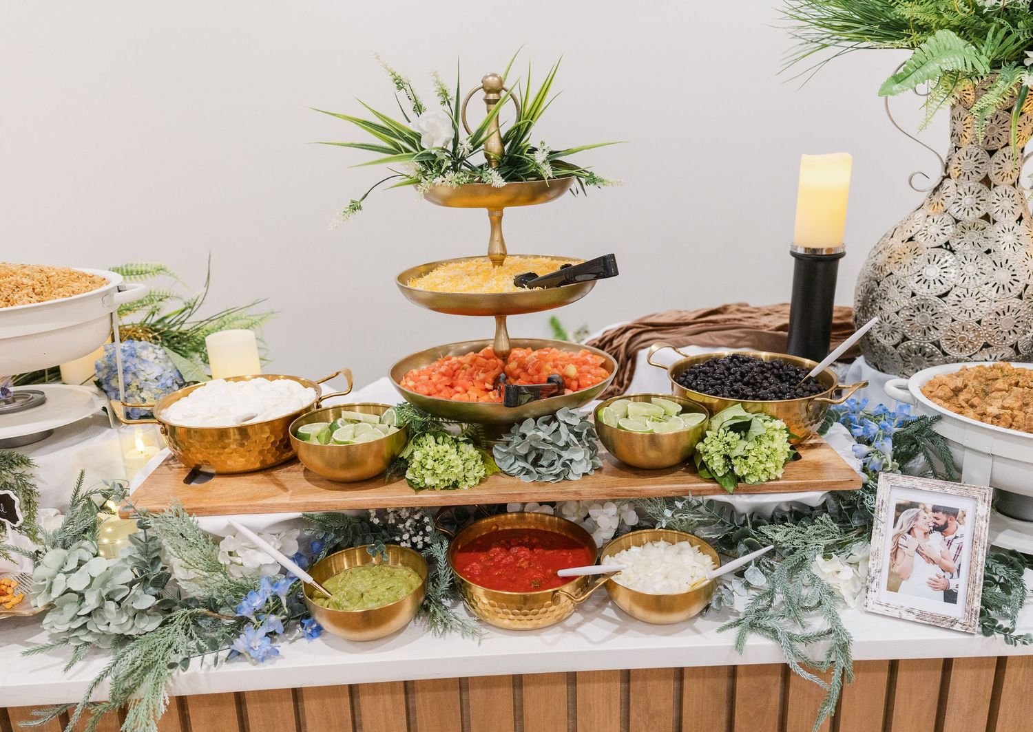 Elegant wedding buffet table with gold bowls, tiered stand, and fresh toppings arranged on a wooden board with greenery.