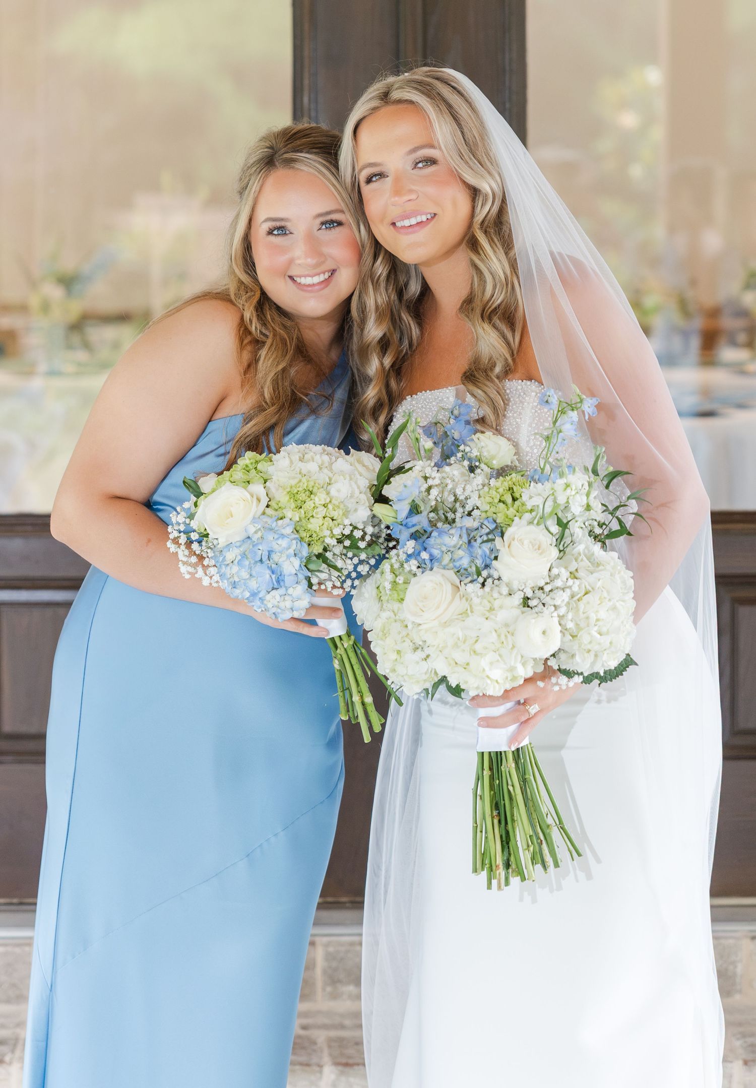 Bride in white gown with veil holding white floral bouquet poses with bridesmaid in light blue dress at wedding reception.