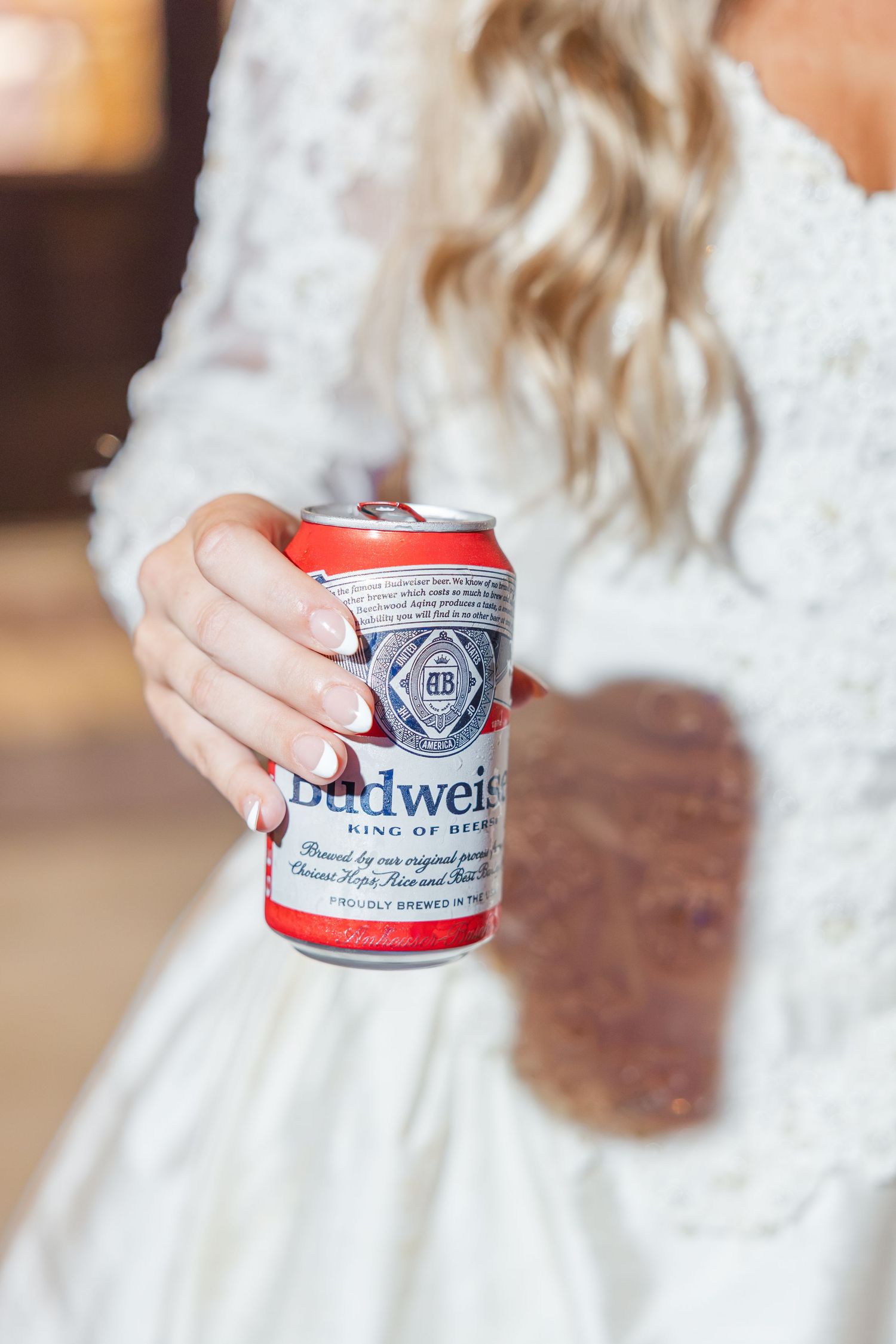 Bride in white dress holding a Budweiser beer can with elegant manicured nails at wedding reception.