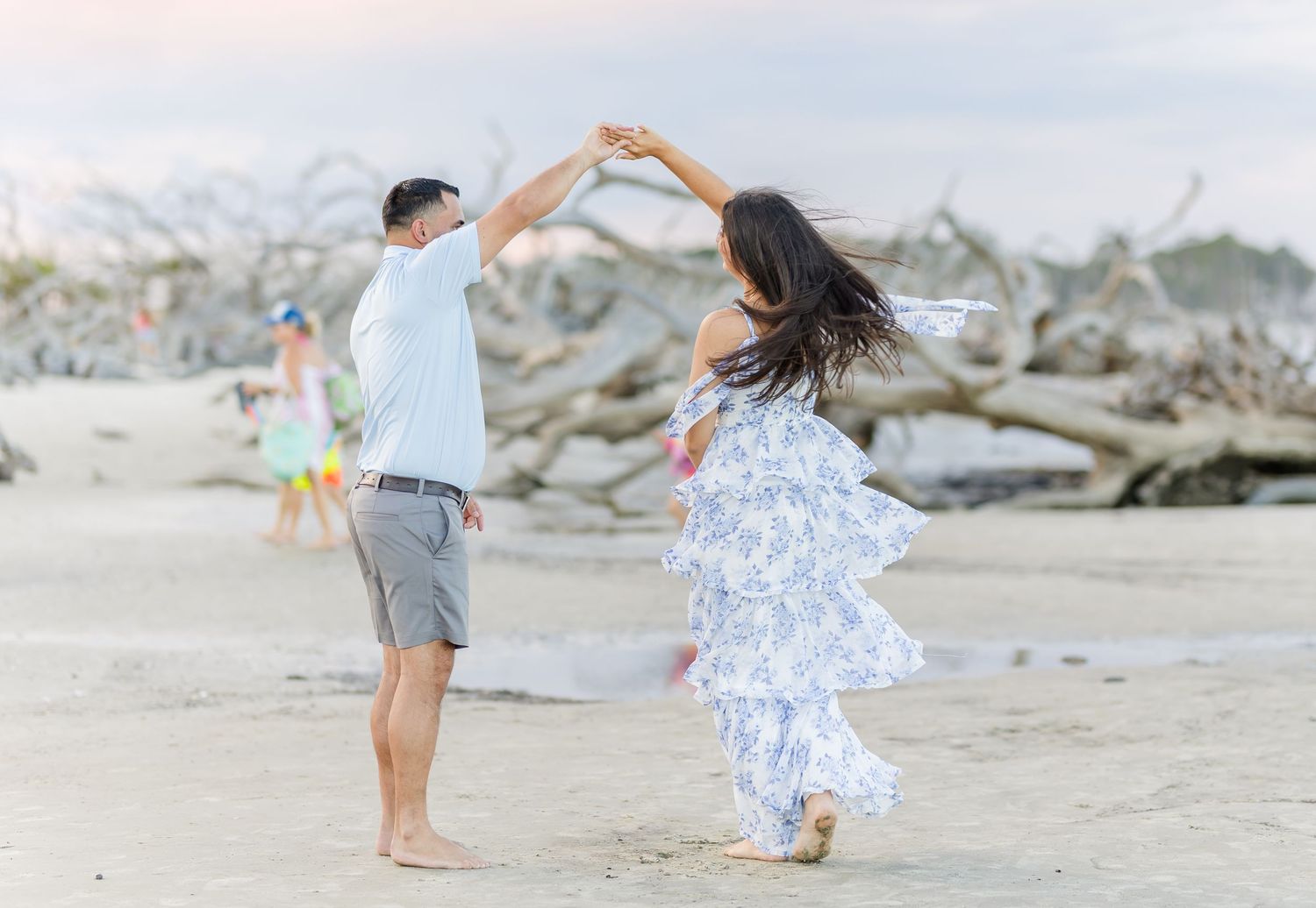 A couple walking on a beach holding hands in romantic poses as their flowy white dress and blue shirt flow in the breeze.