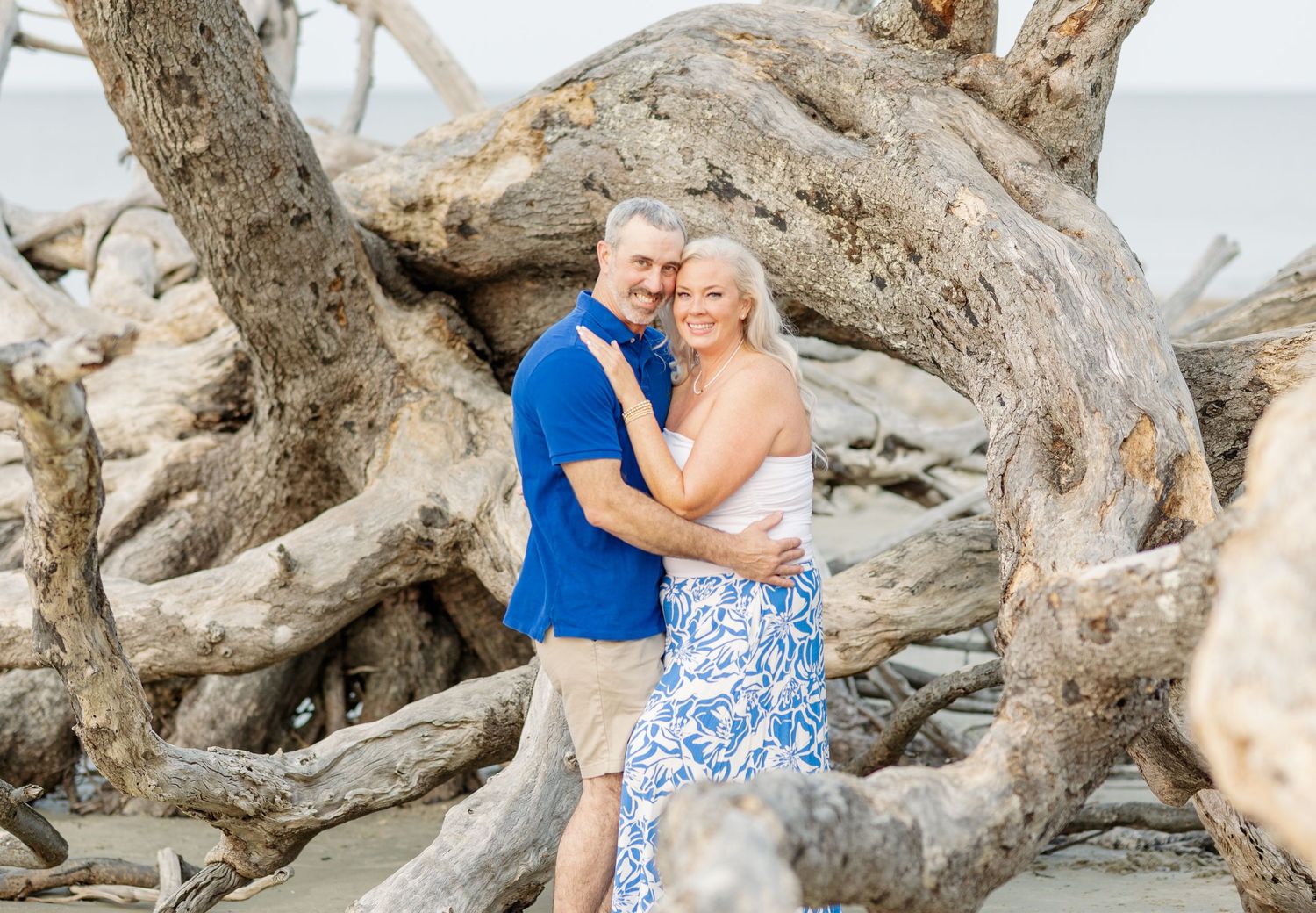 A couple poses together on a beach among weathered driftwood logs and rocks during a warm sunny day.