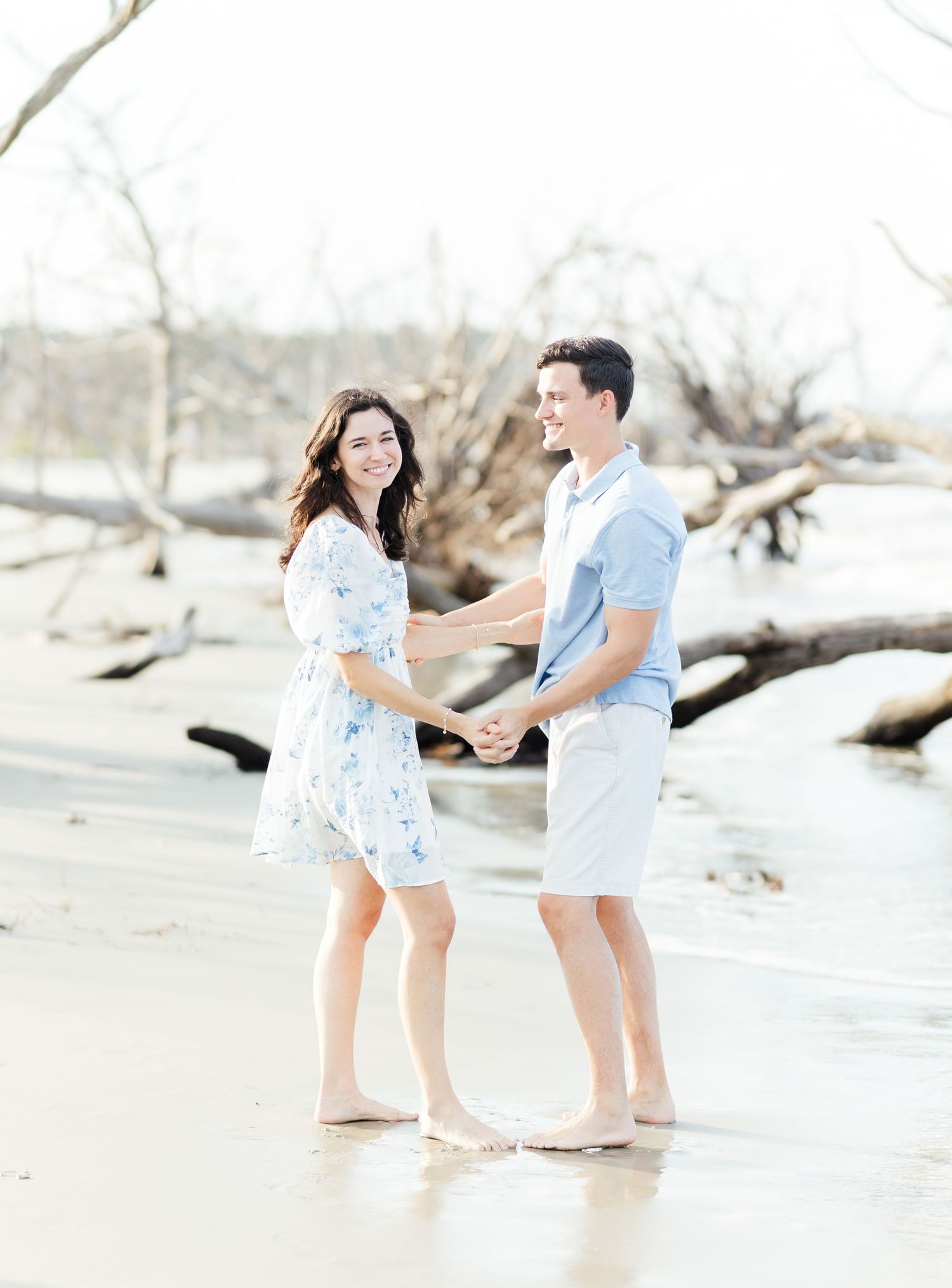 Couple holding hands on a bright, dreamy beach with driftwood, wearing light blue and white casual outfits.