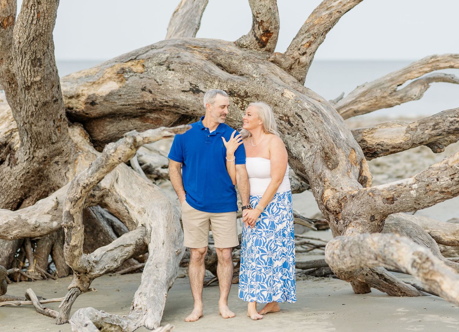 A couple stands together on a beach among large driftwood logs and branches in casual summer attire.