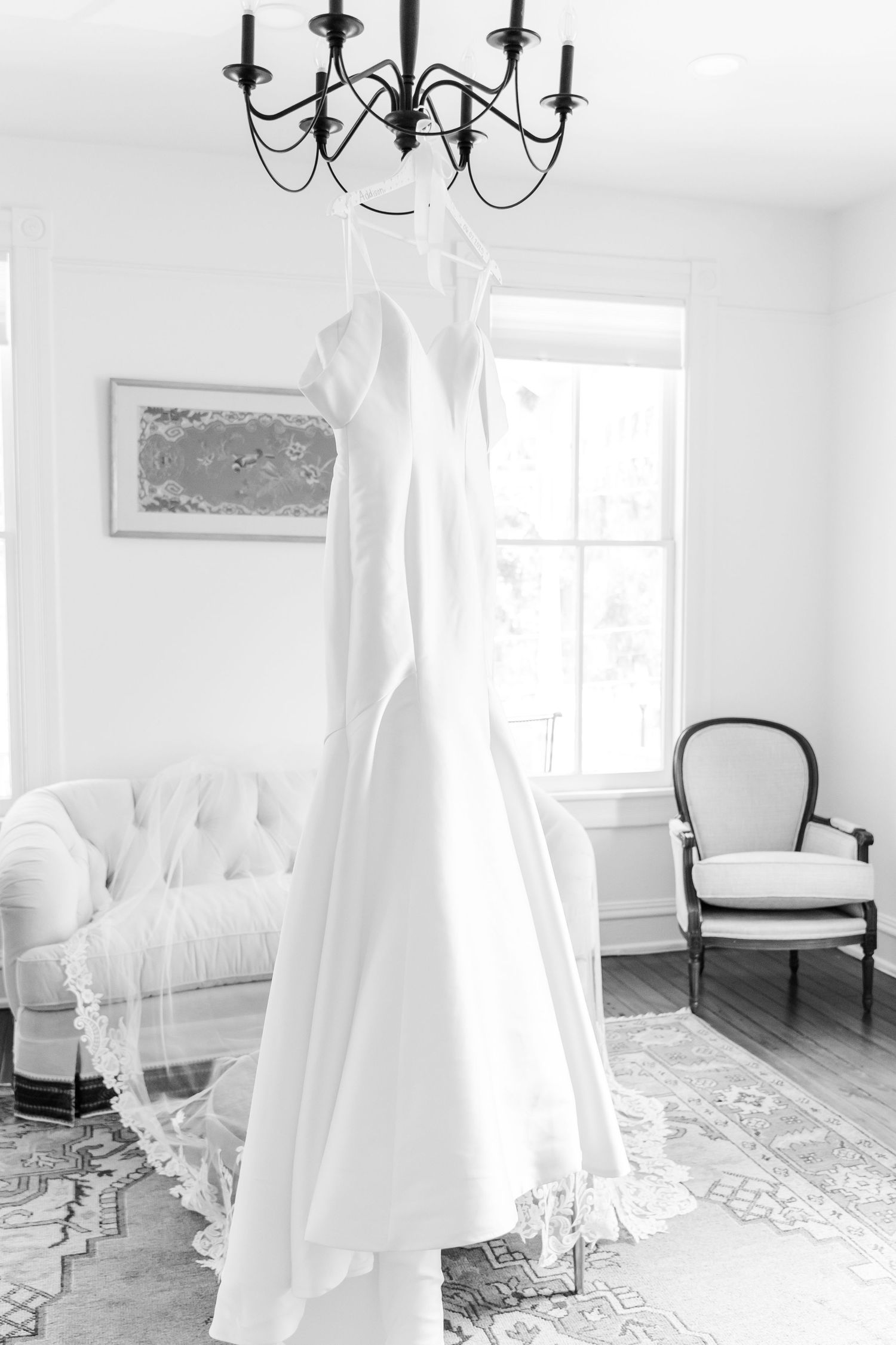 Sleek white wedding dress hanging from black chandelier in airy white room with vintage furniture.