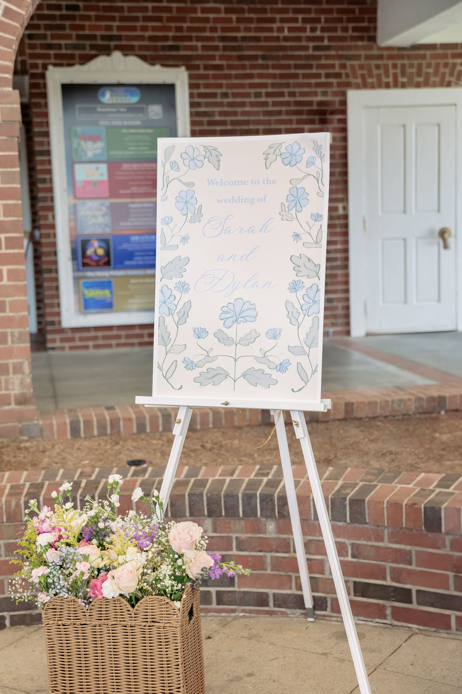 Decorative welcome sign on white easel with blue floral design next to basket of pink flowers by brick entrance.