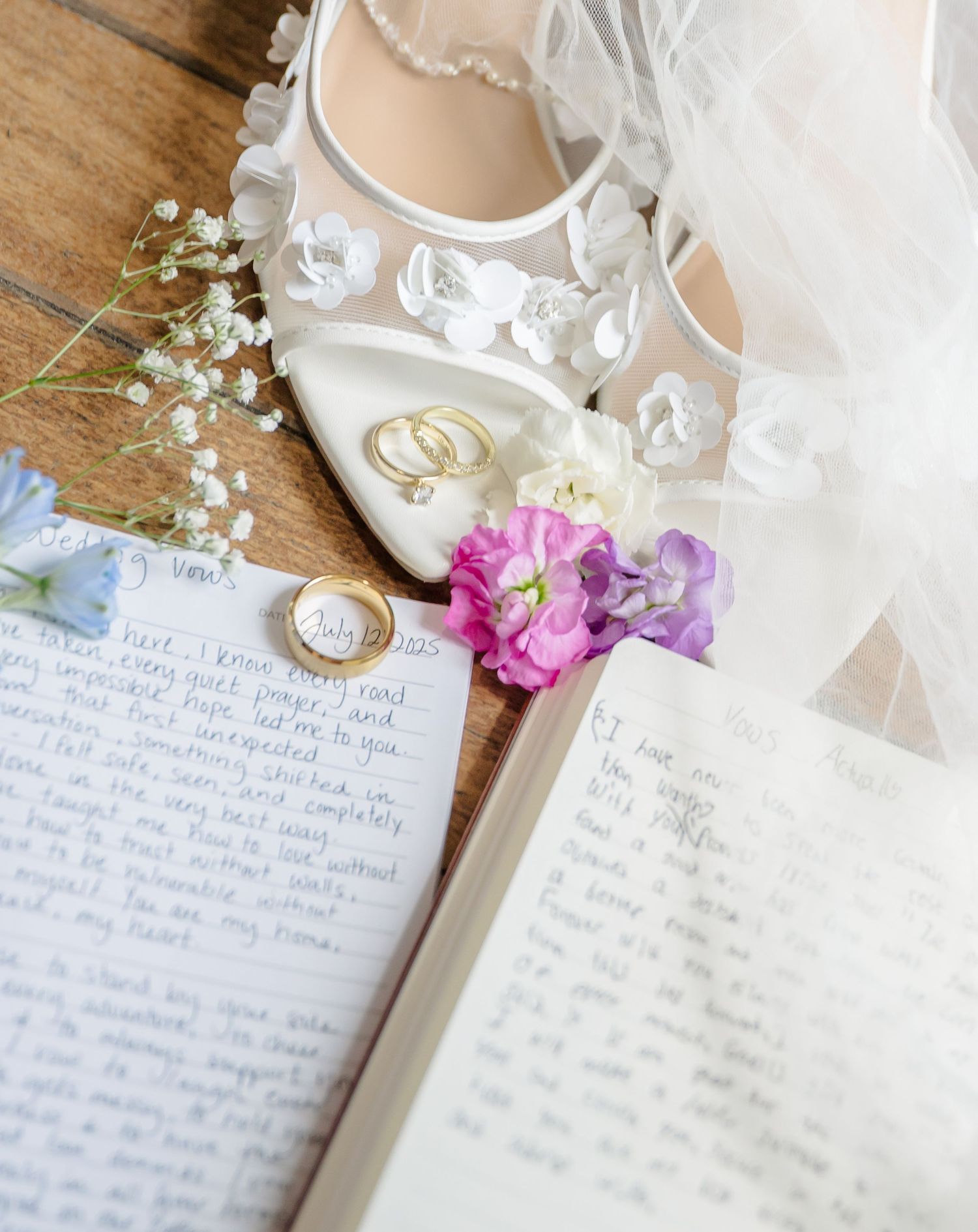Wedding details including a white bridal shoe, delicate veil, handwritten vows, and floral accents arranged on wooden surface.