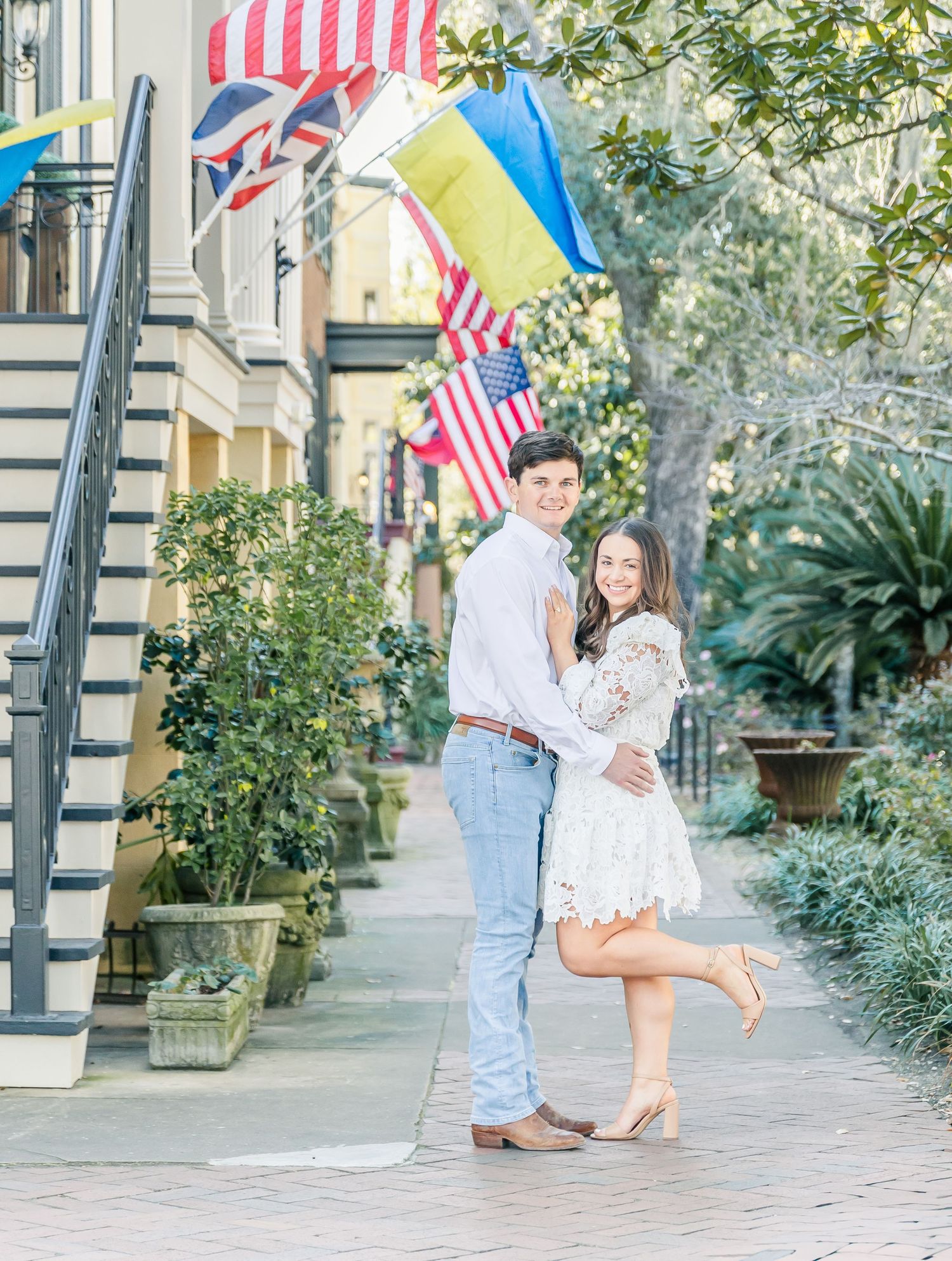 A couple poses on a brick sidewalk in downtown Charleston with American flags and greenery in the background.
