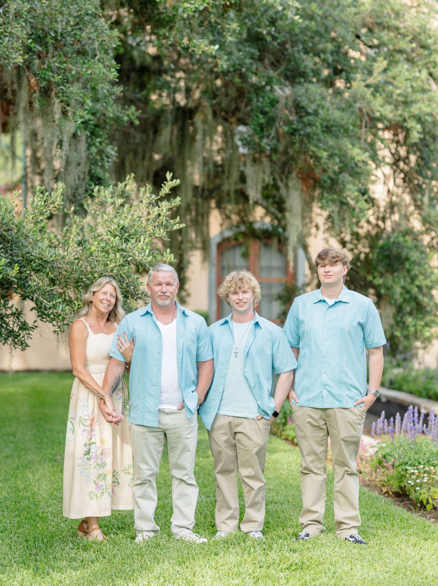 A family wearing coordinated light blue and beige outfits poses together on grass with Spanish moss trees in background.
