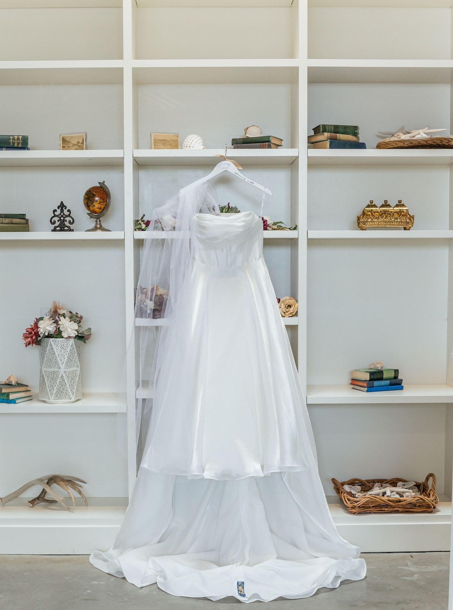 White wedding dress displayed on hanger against built-in bookshelves decorated with vintage accessories.