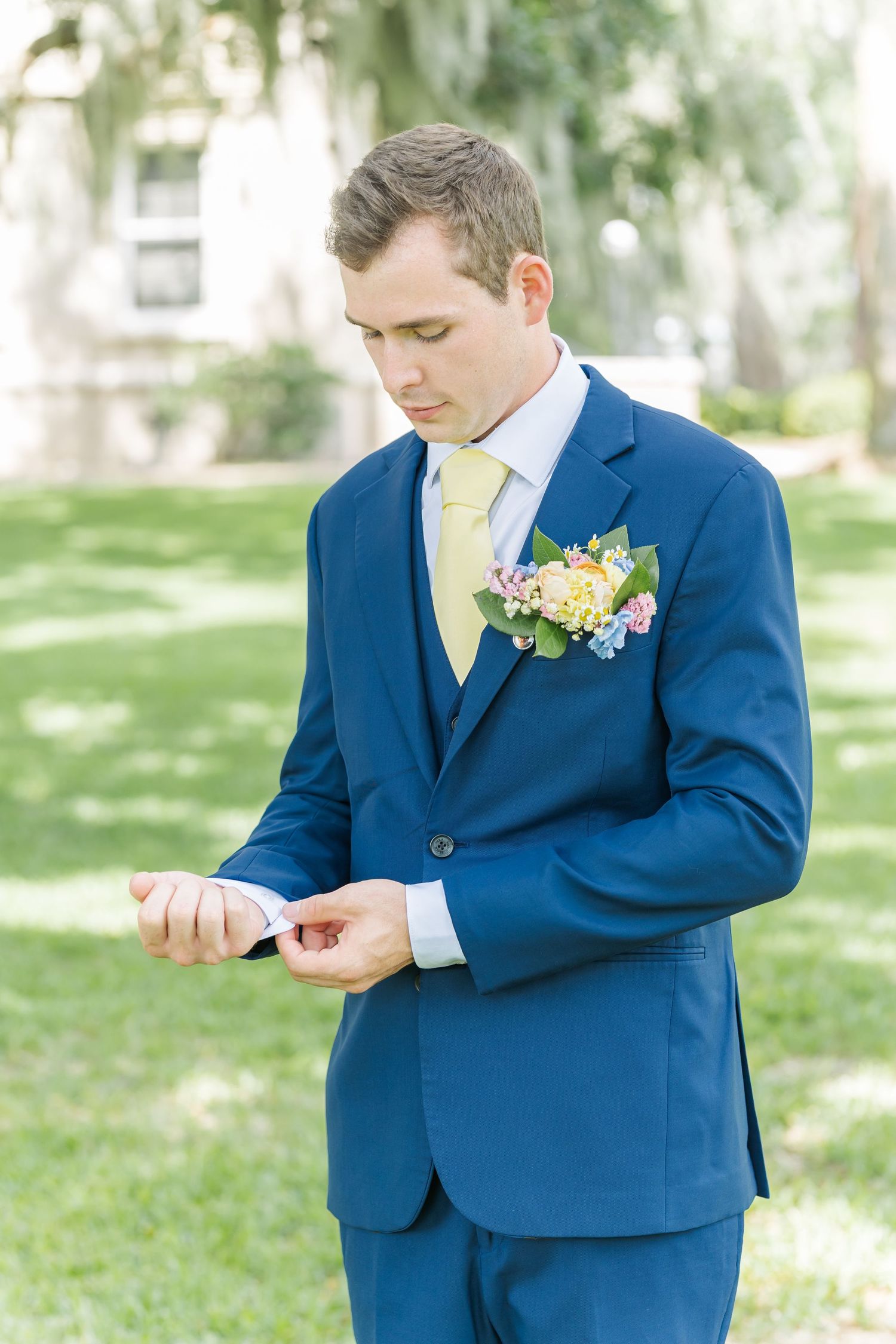 Groom in bright blue suit with yellow boutonniere looks at wedding program in outdoor garden setting.