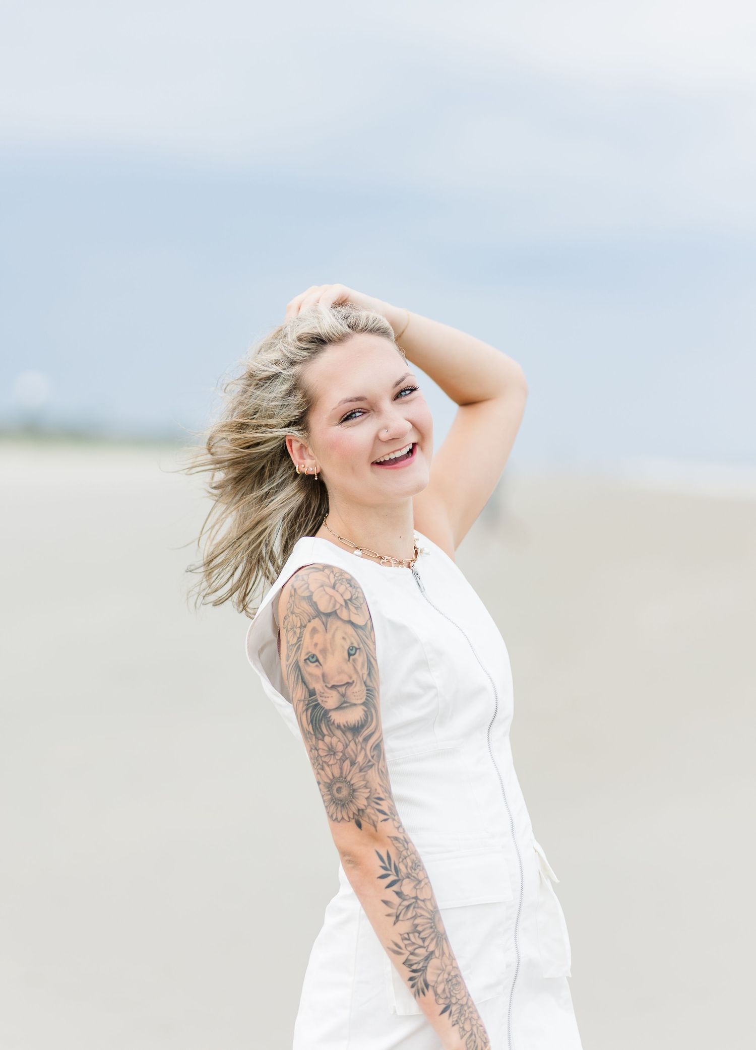Candid beachside moment captured with wind-swept hair against soft blue sky backdrop.