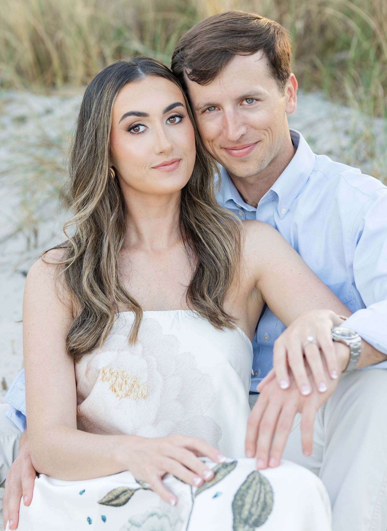 Couple sitting together on sandy beach dunes, woman in floral dress with long wavy hair, man in light blue shirt smiling.