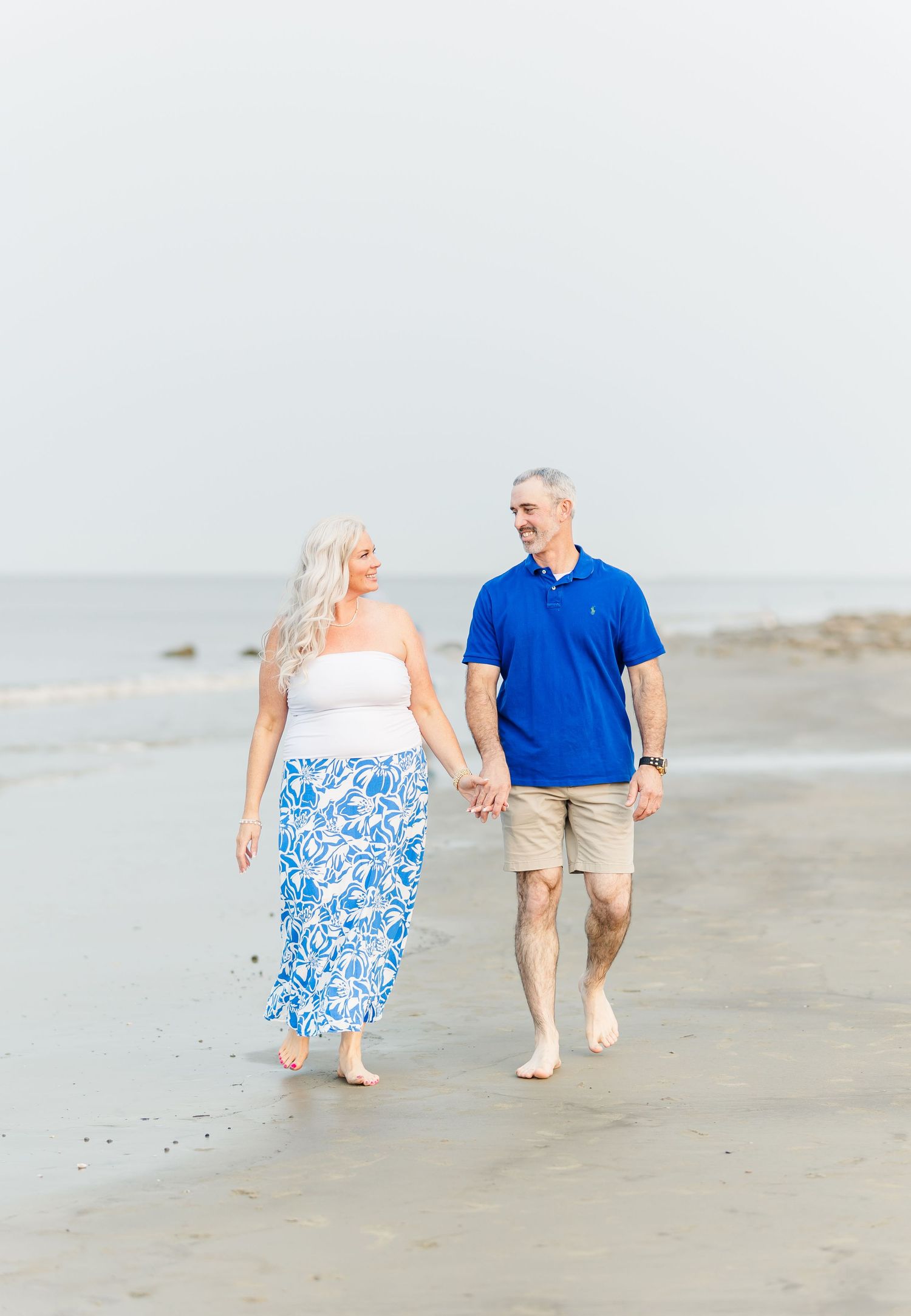 Couple holding hands while walking barefoot along a misty beach in coordinating blue and white outfits.