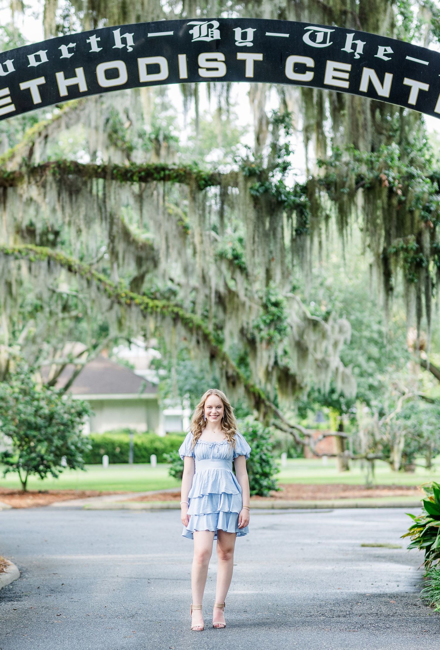 A person in a blue tiered dress stands under a Methodist Center archway with Spanish moss draped trees.