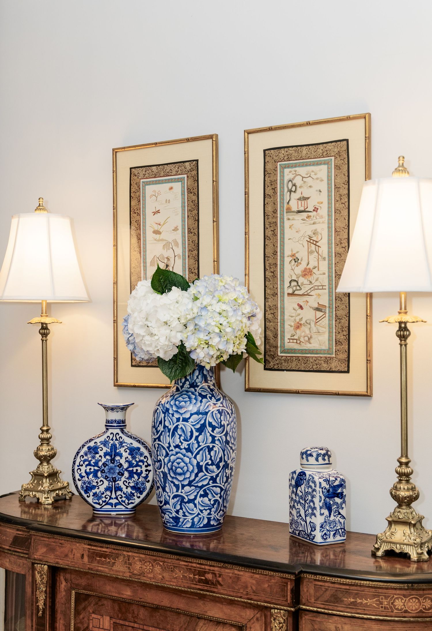 Blue and white chinoiserie vases with white hydrangeas on wooden sideboard flanked by gold lamps and framed artwork.