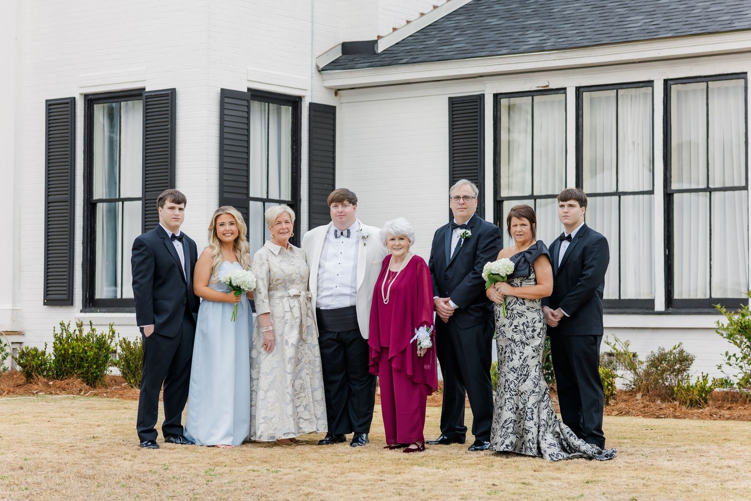 Wedding family portrait group of eight posed outside white venue with black shuttered windows.