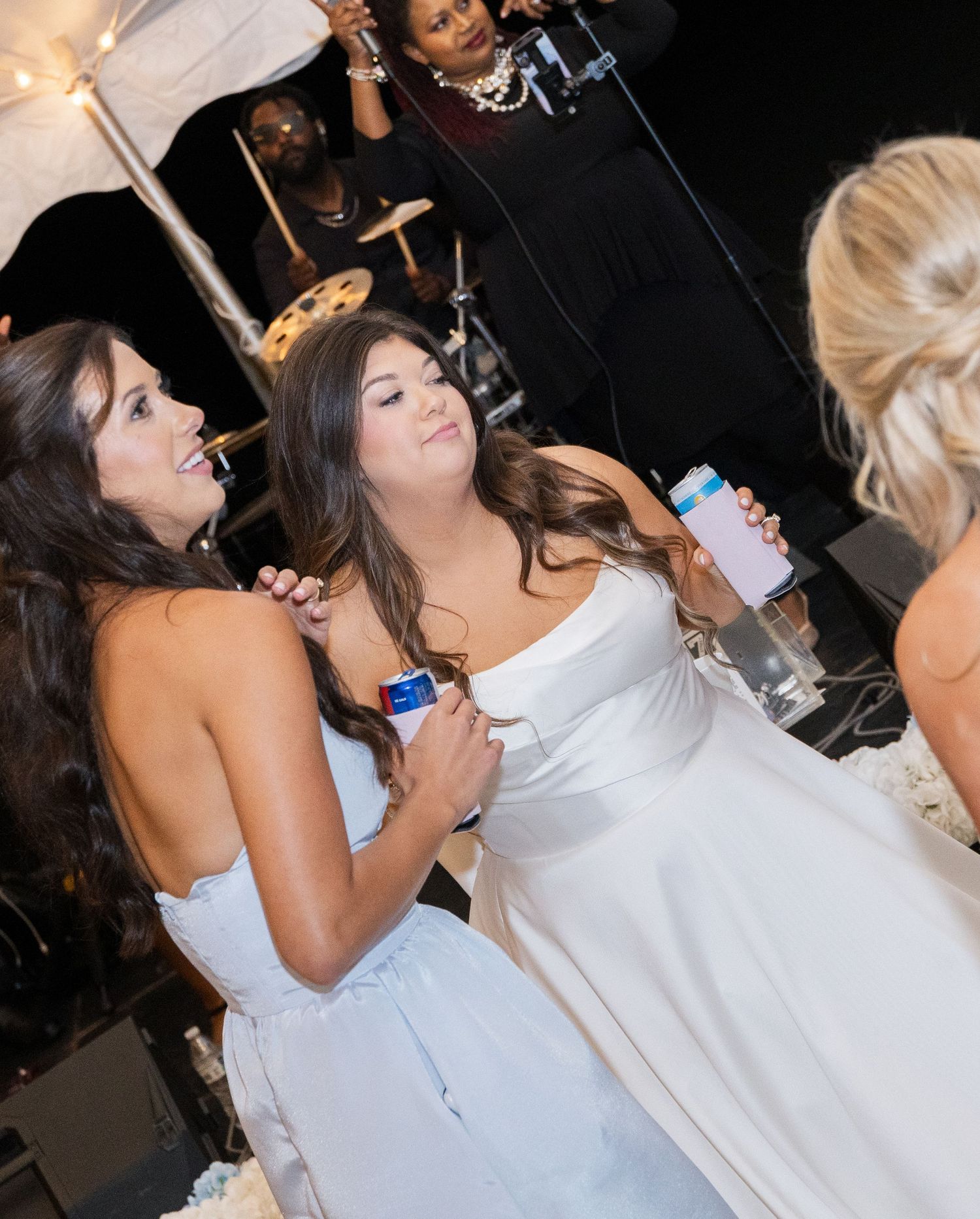 Two women in white dresses chat and hold drinks at a wedding reception with a band performing in the background.