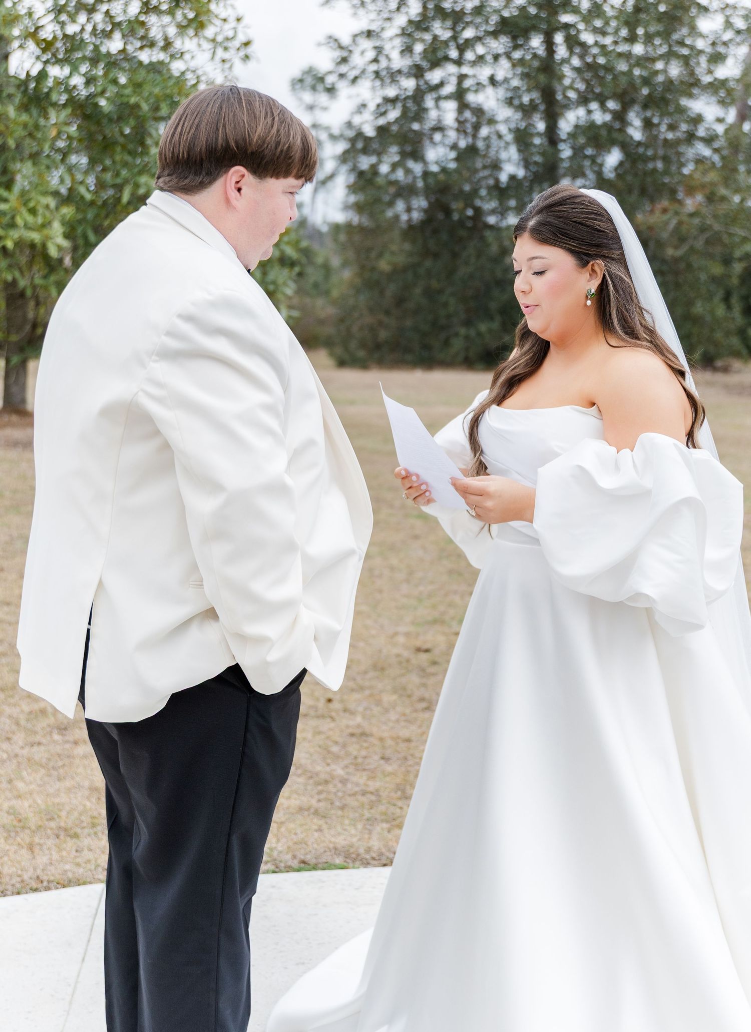 Bride in strapless gown with puff sleeves reads vows to groom in white jacket during outdoor first look.