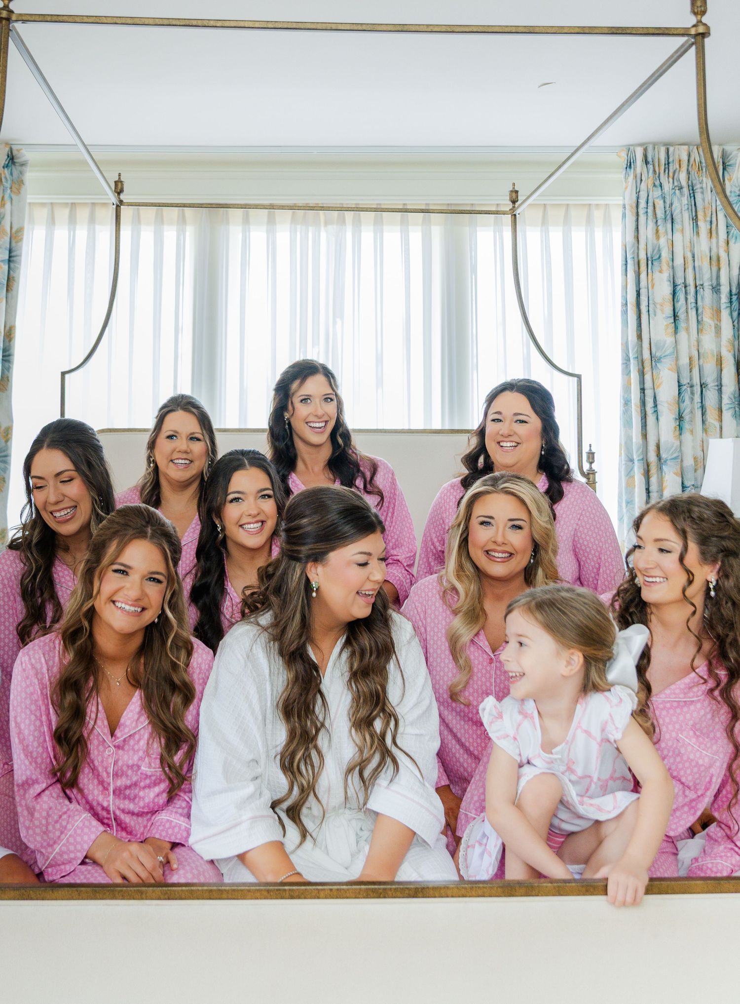Bride in white robe surrounded by bridesmaids in pink pajamas sitting on a canopy bed during wedding morning preparations.