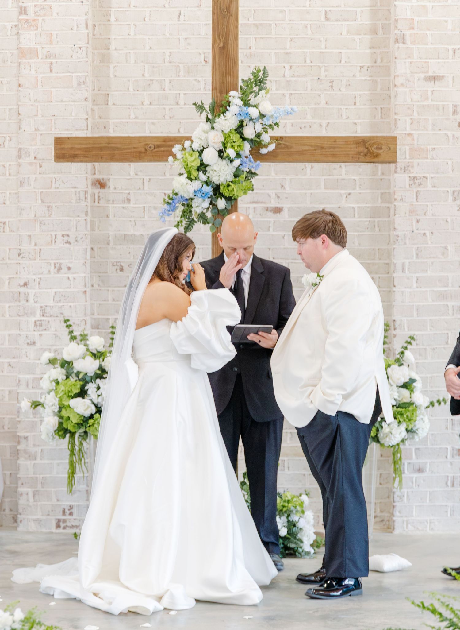 Bride and groom exchange vows beneath wooden cross adorned with blue and white flowers at altar.