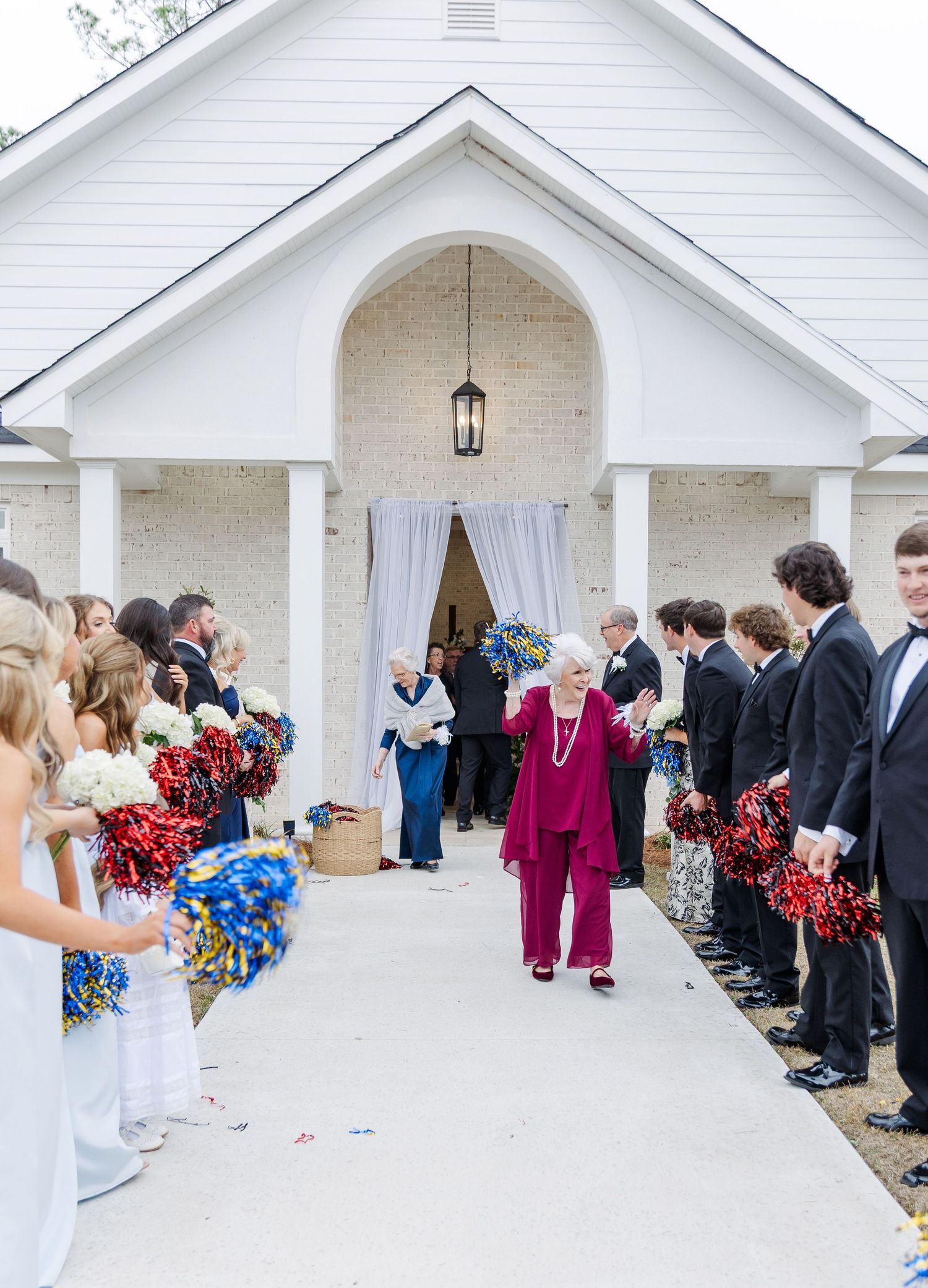 Wedding guests form celebratory tunnel with colorful pom-poms outside white brick chapel entrance.