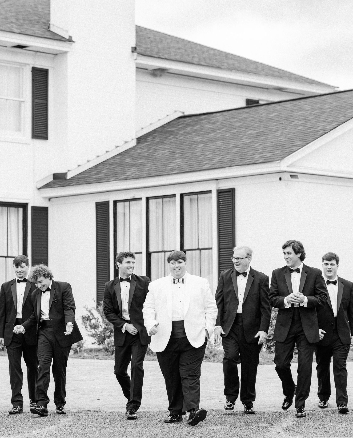 Black and white photo of groom in white tuxedo walking with groomsmen in black tuxedos outside venue.