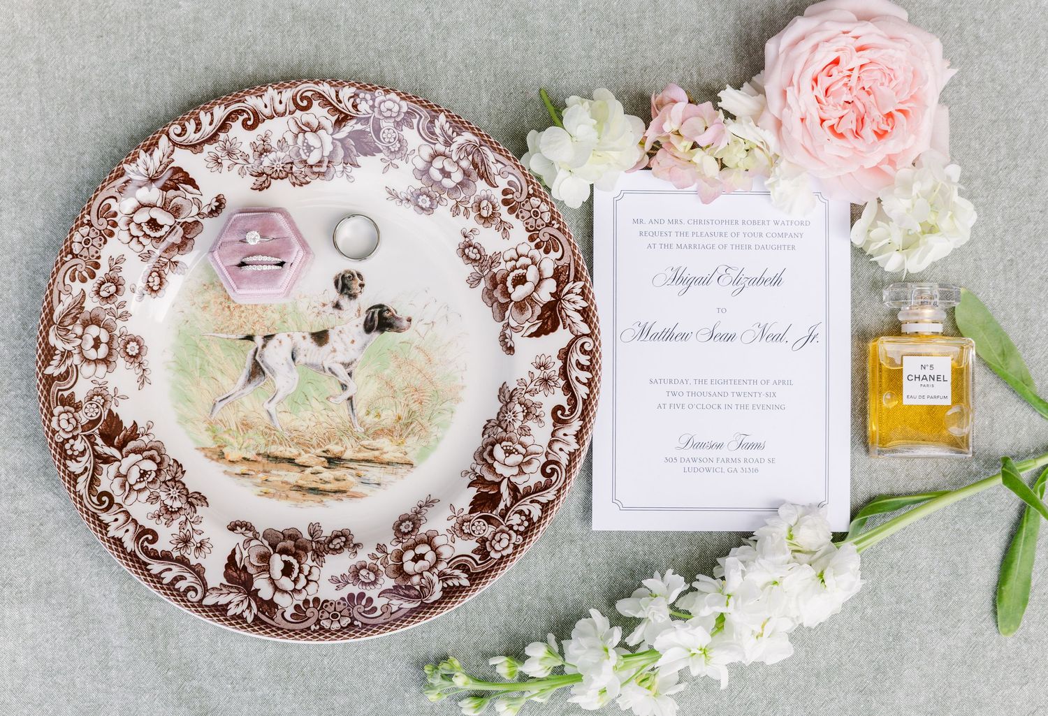Elegant wedding flat lay with ornate brown transferware plate, rings, invitation, Chanel perfume, and pink florals.